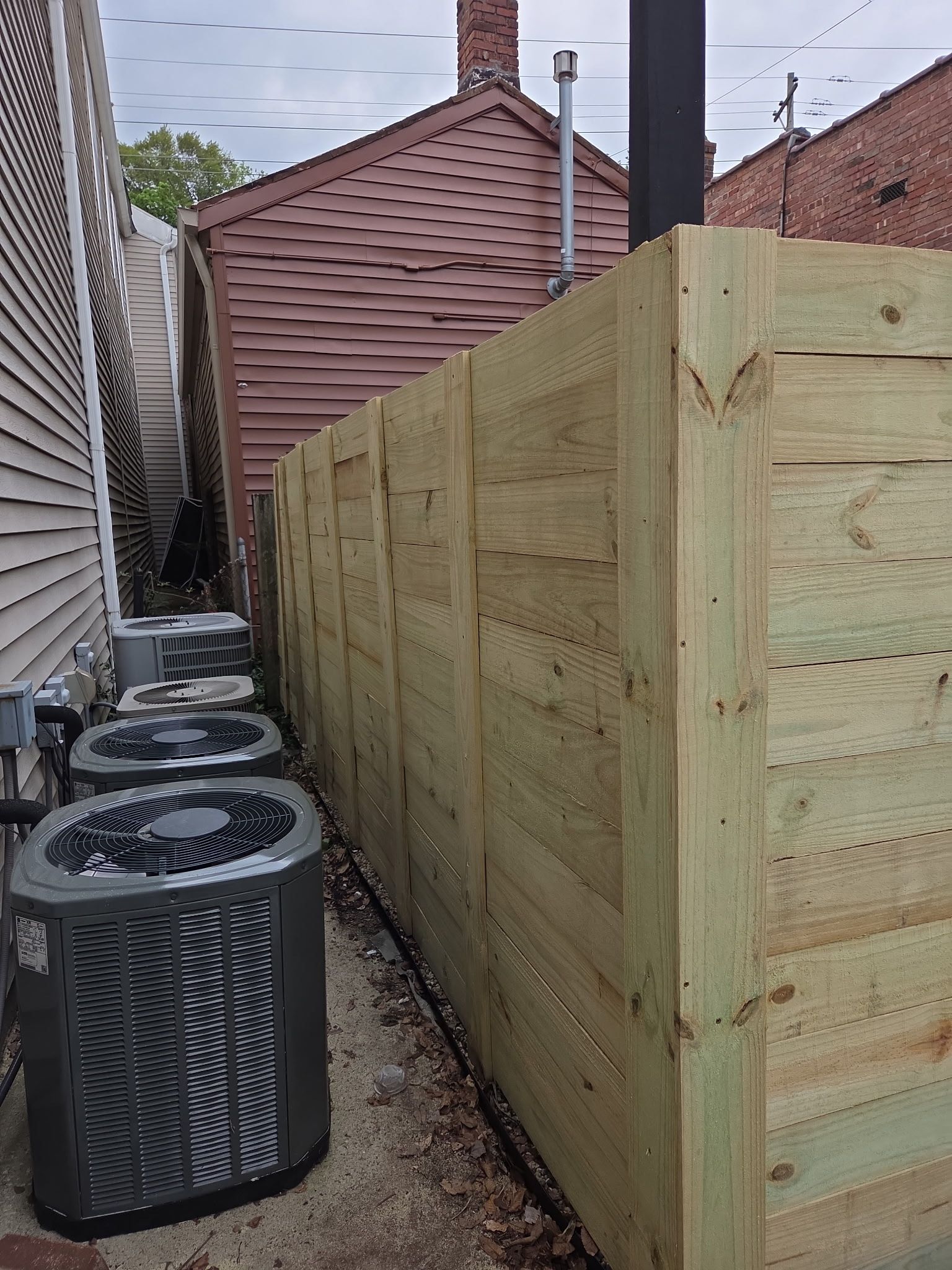 A new light-wood fence stands in a narrow alleyway next to three gray HVAC units placed against a building wall.