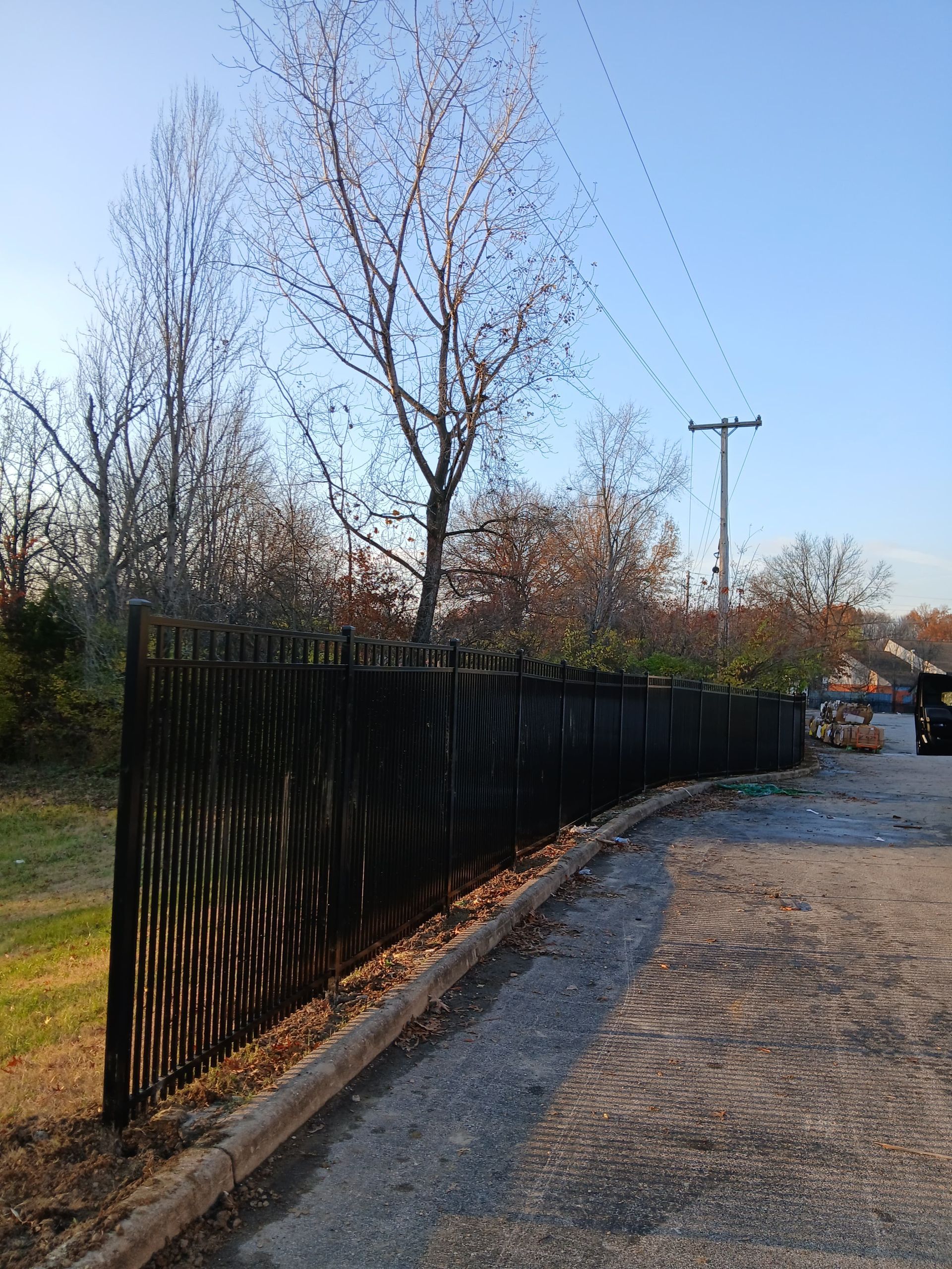 Black wooden fence along a gravel road, trees in the background, power lines overhead.
