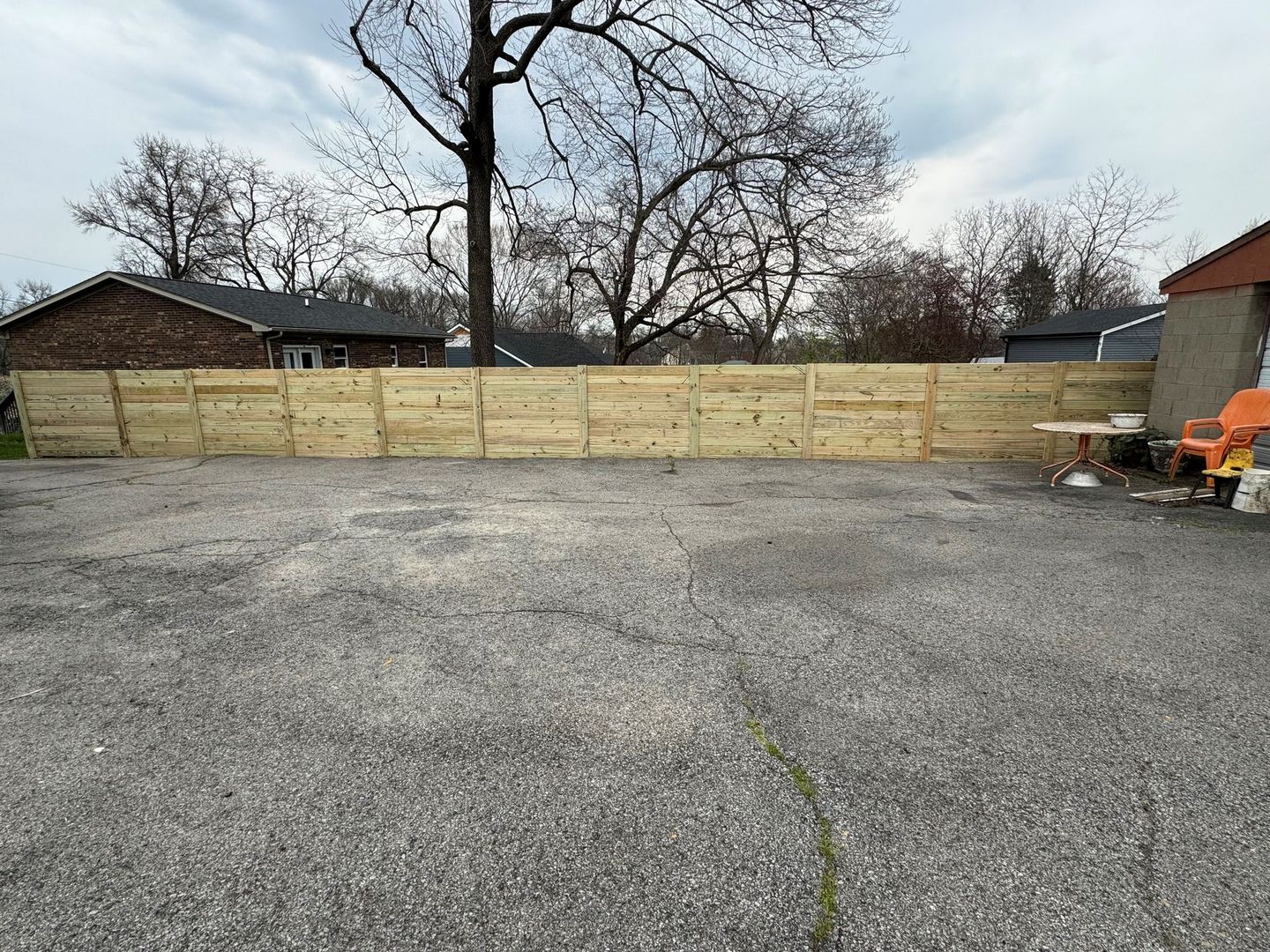 Empty gravel lot with a wooden fence, leafless trees, and a small building on the right.