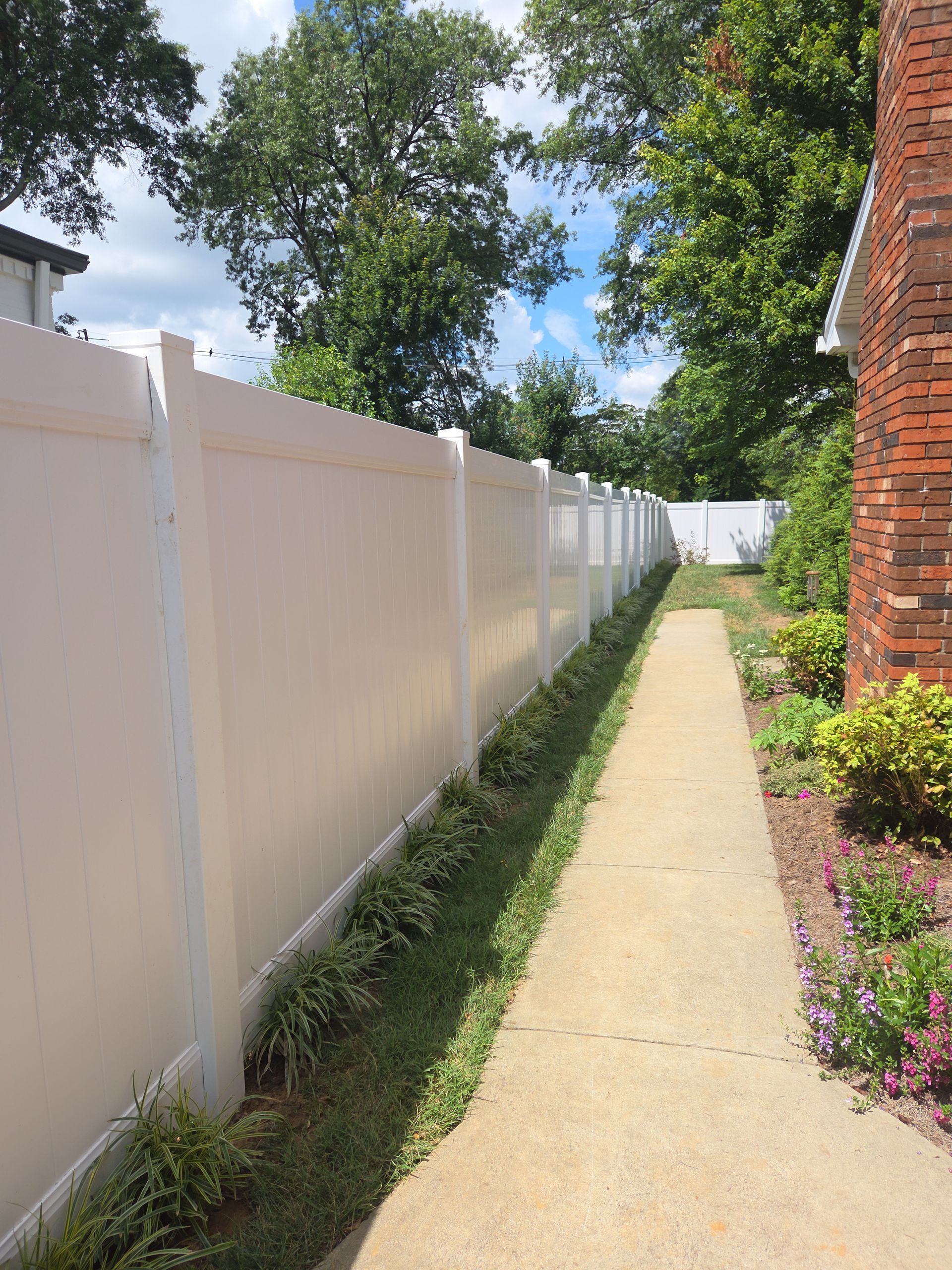 White fence alongside a concrete path, next to a brick building and green foliage under a blue sky.