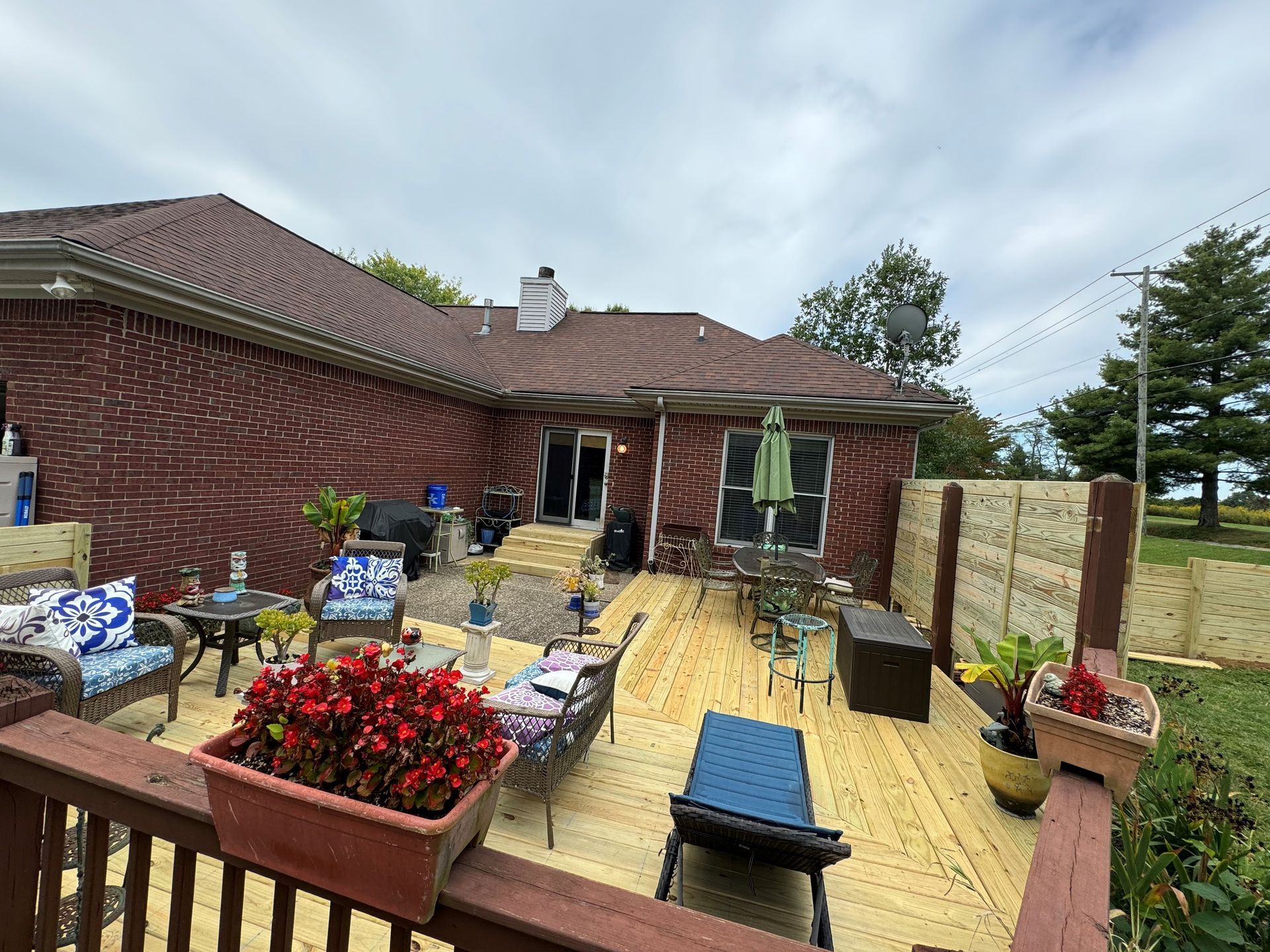 Backyard patio with wooden deck, red flower planters, outdoor furniture, and a brick house under cloudy skies