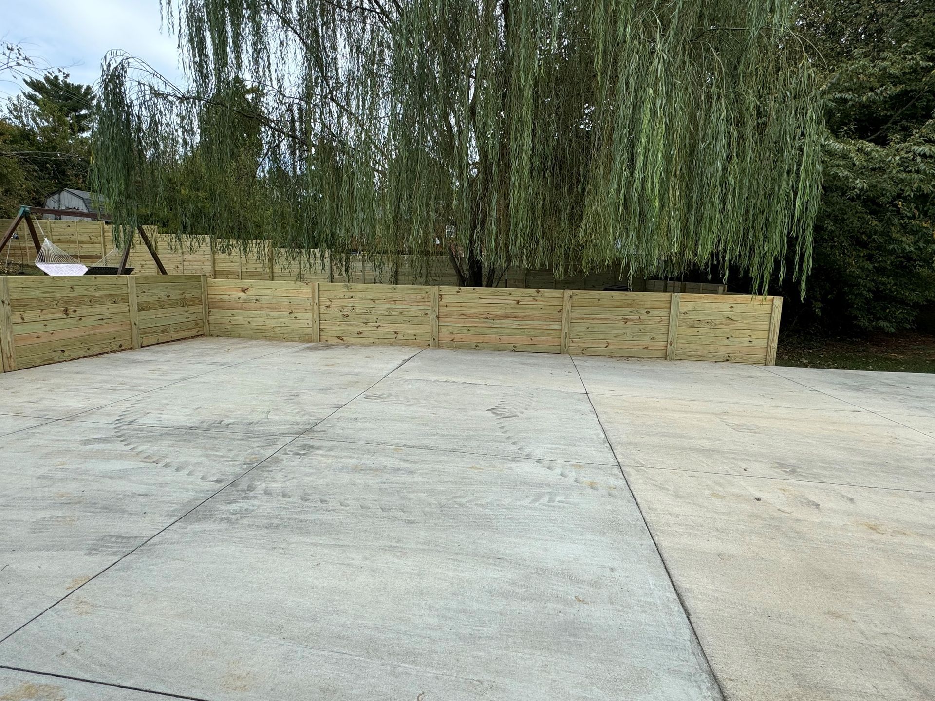 Empty concrete patio with a wooden privacy fence and drooping willow trees in the background