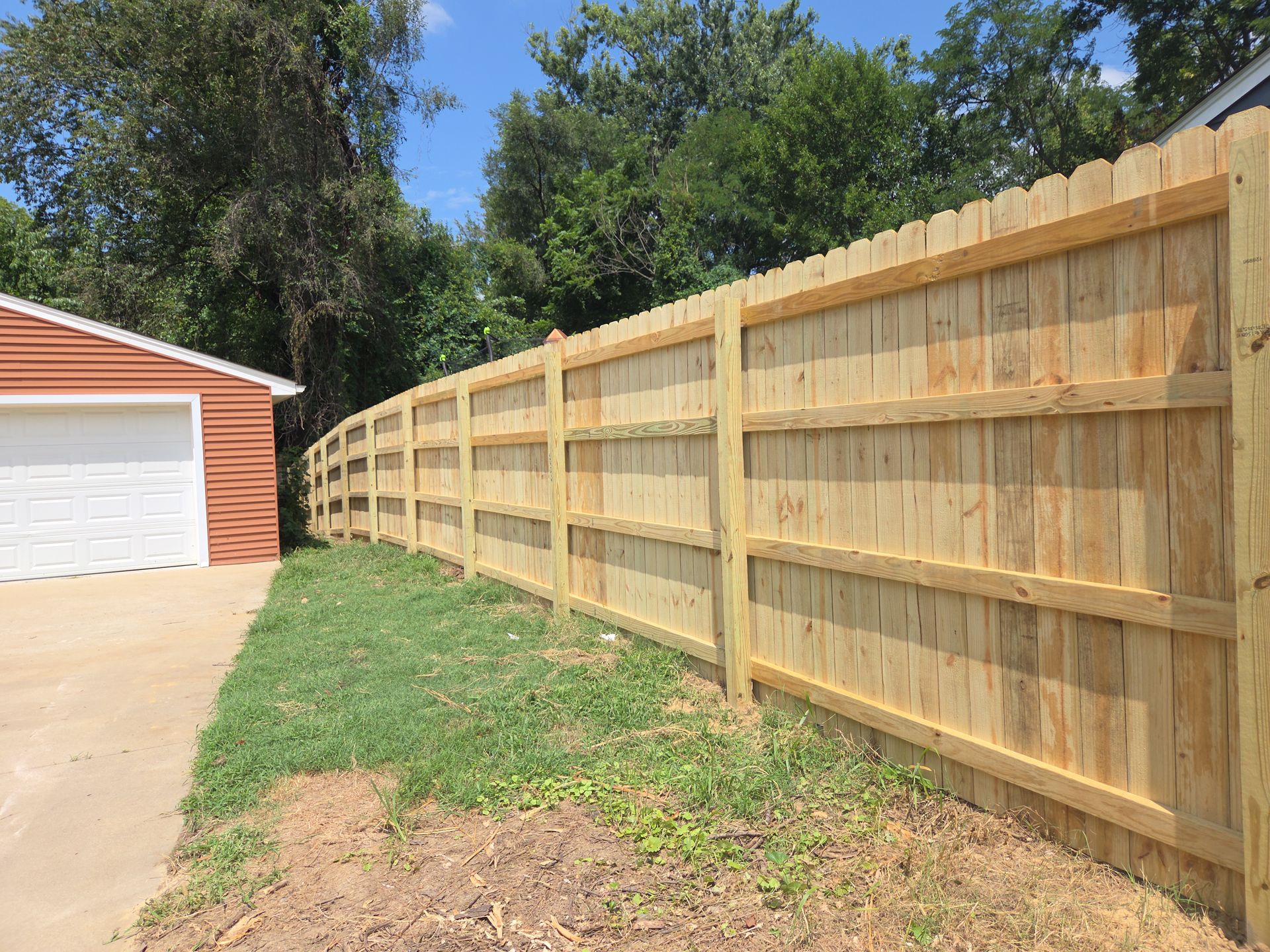 Long wooden privacy fence beside a driveway, with a brick garage and trees under a blue sky