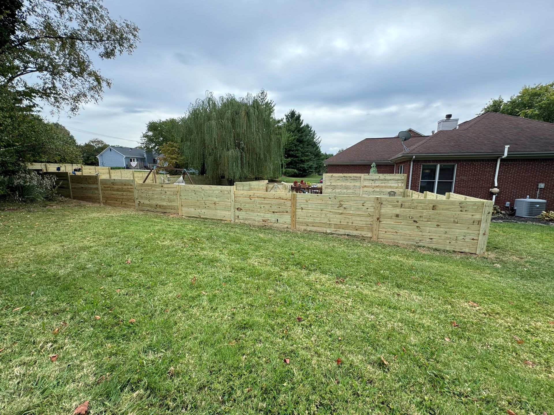 Backyard with grass, a low wooden fence, trees, and a brick house under cloudy skies