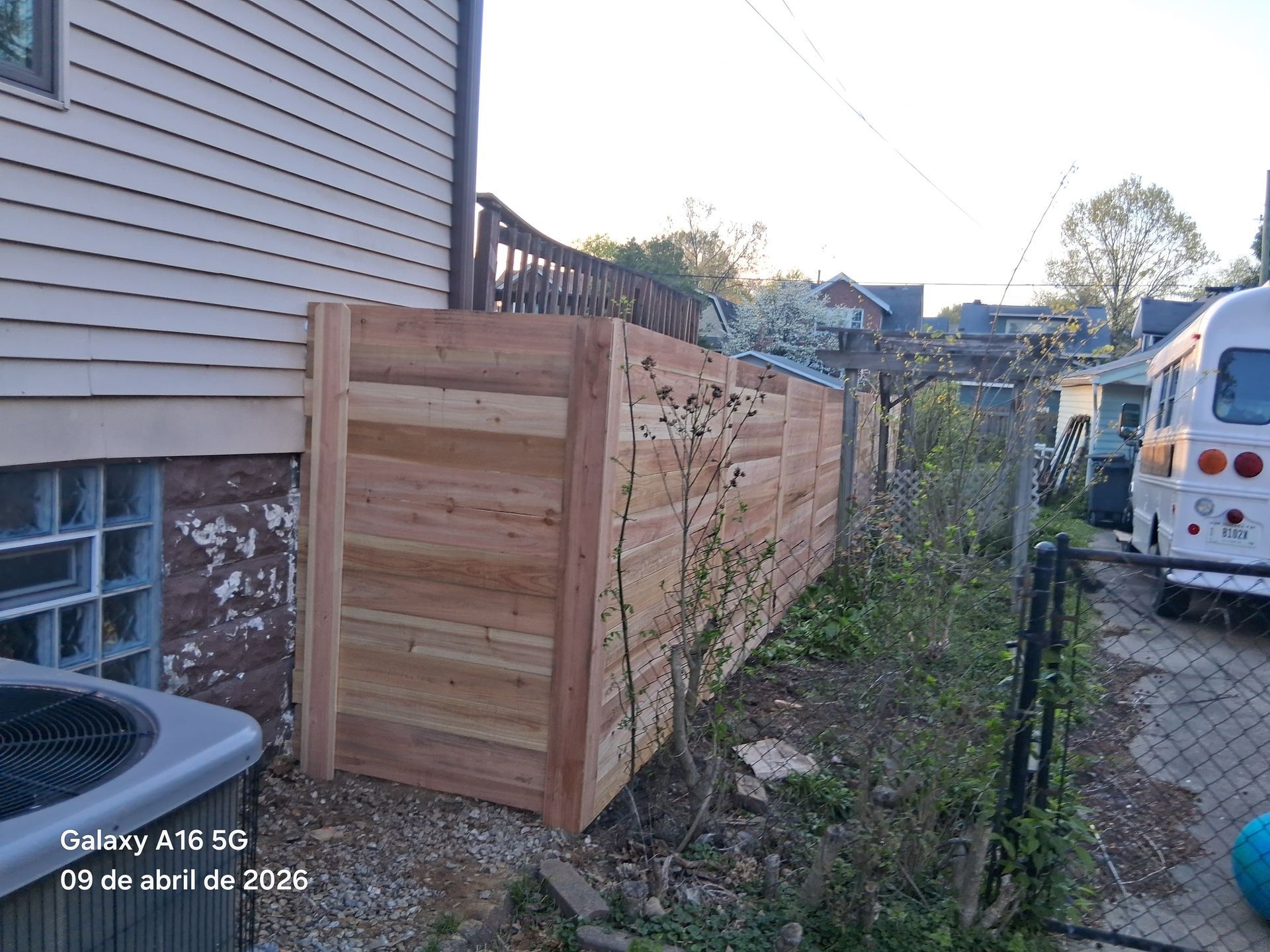 A new horizontal wooden fence stands next to a house exterior, adjacent to a small yard and a white vehicle.
