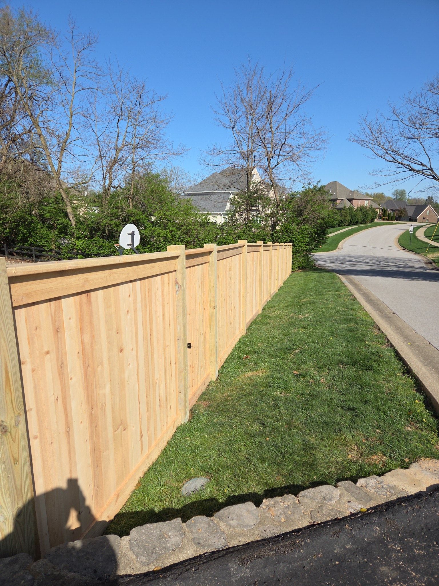 Wooden privacy fence along a grassy strip beside a curving residential street on a sunny day