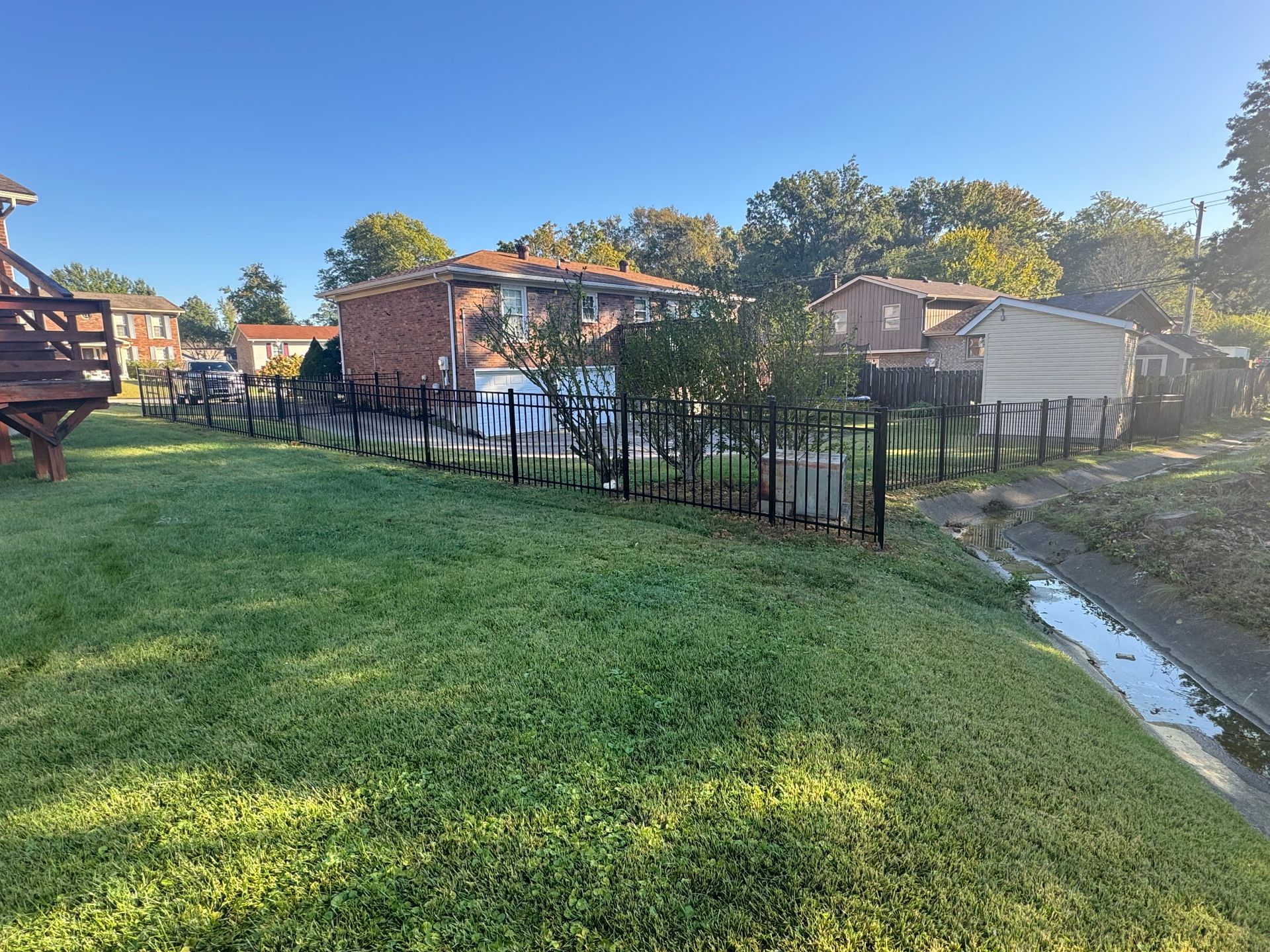 Backyard with grass, black fence, small sheds, and a narrow water-filled ditch under a clear blue sky