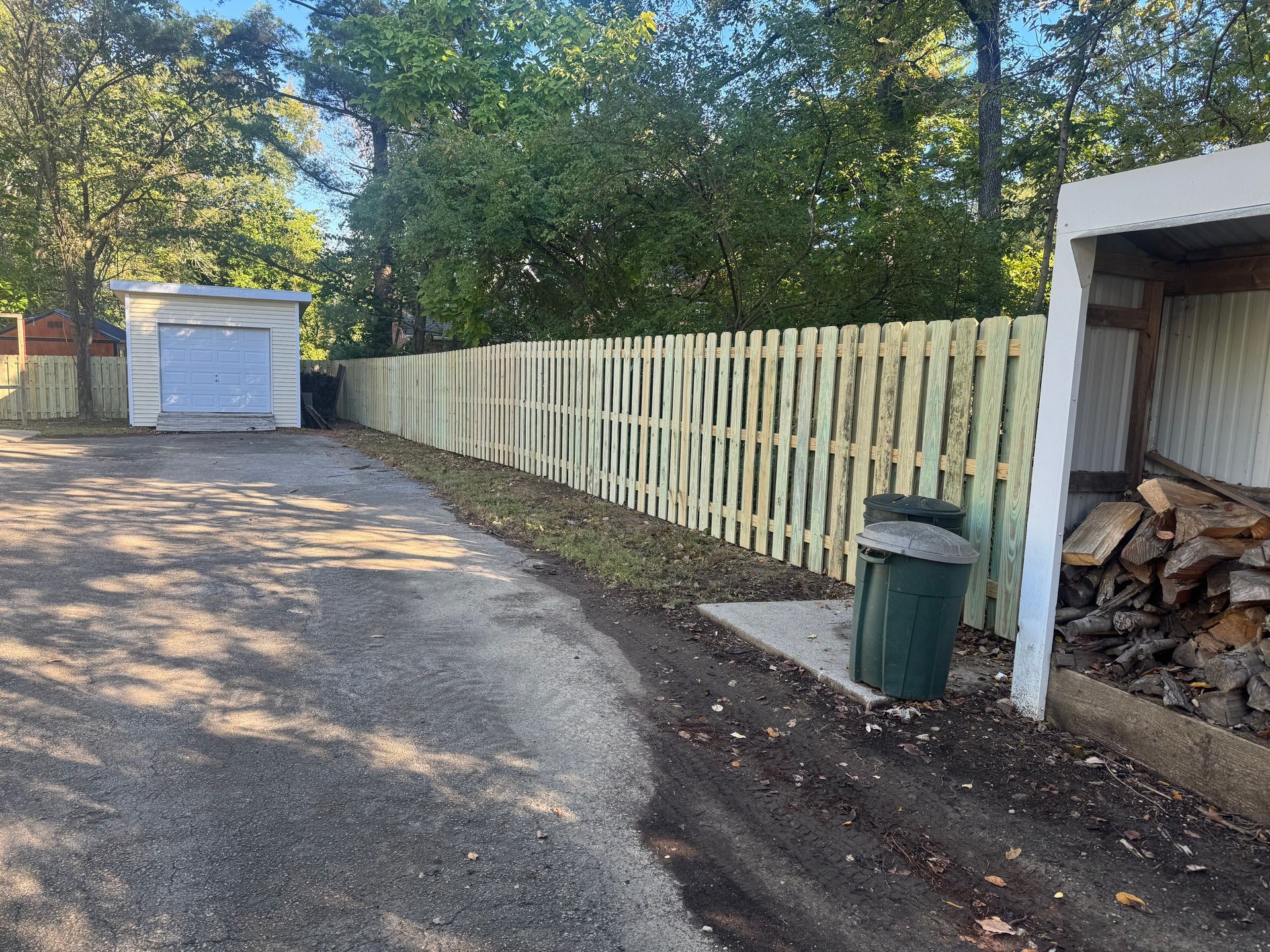 Gravel driveway beside a white fence, green trash bin, and wood shed with stacked firewood.