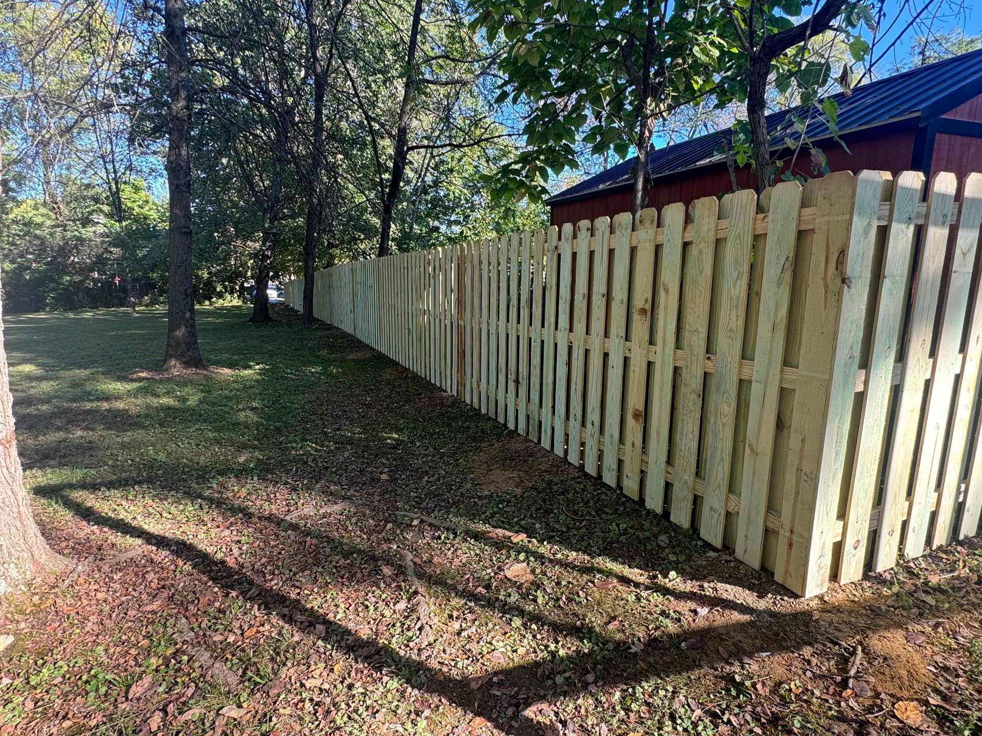 Wooden fence along a shaded grassy yard beside trees and a house with a blue roof