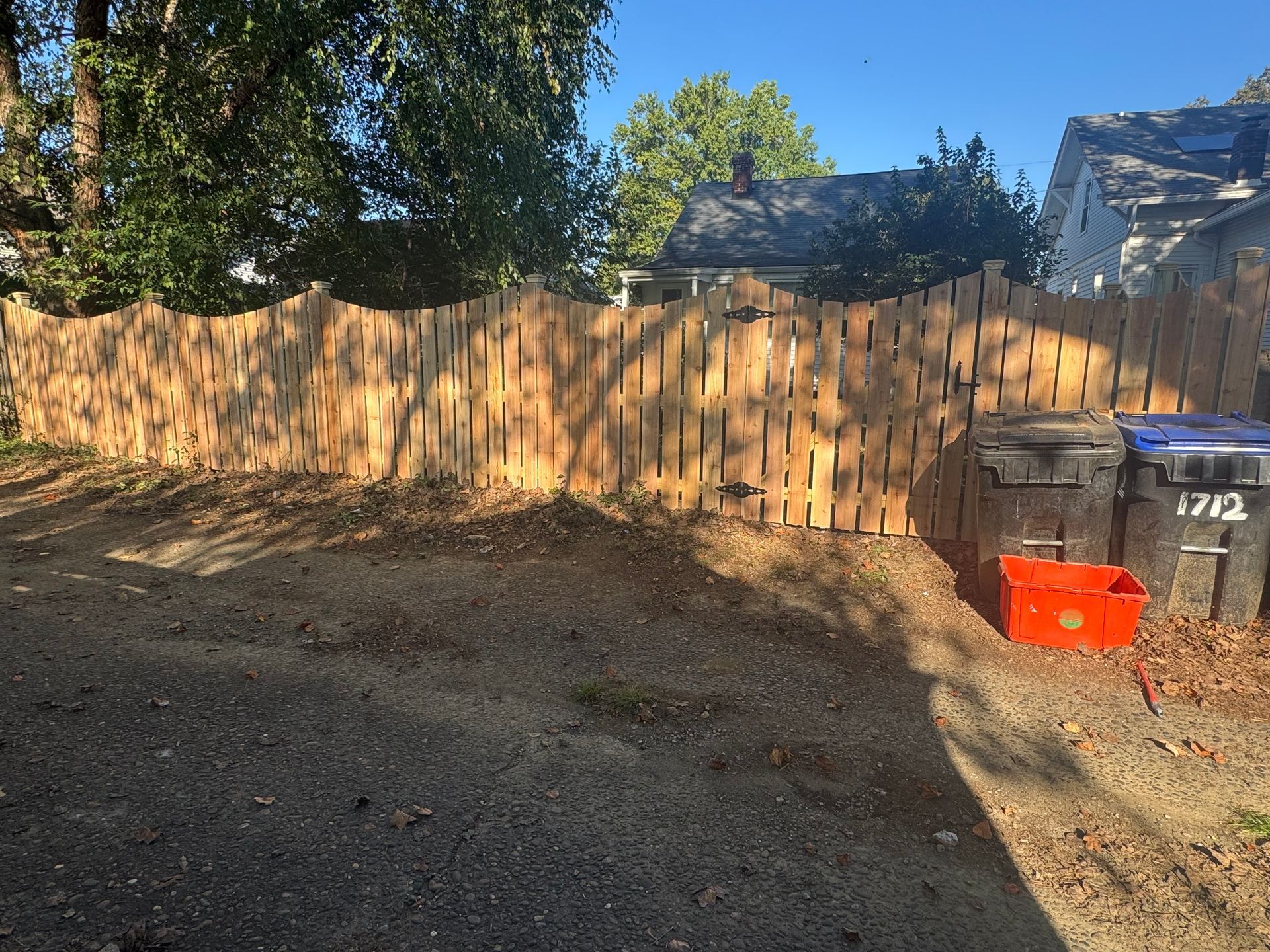 Wooden backyard fence in sunlight, with trash bins and a red bin beside a gravel driveway.
