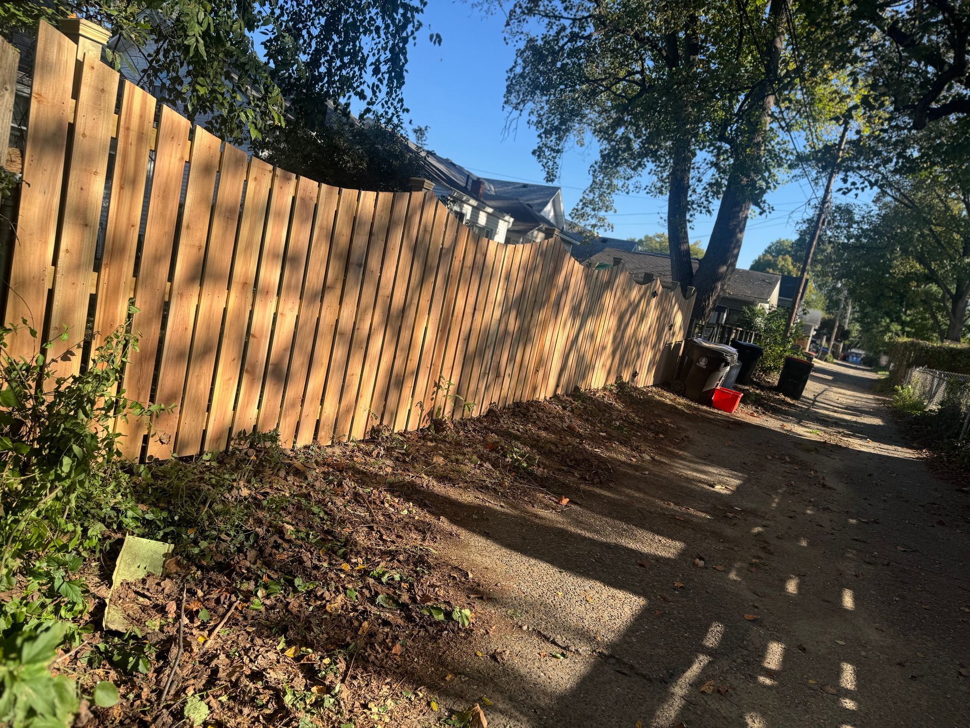 Sunlit dirt path beside a wooden fence, with trees casting shadows and a red bucket near the end.