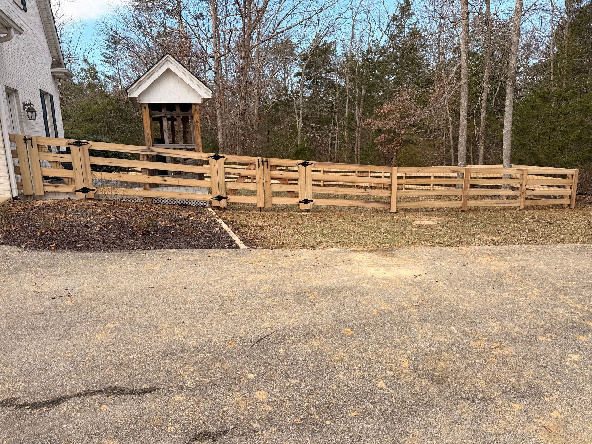 Wooden fence surrounding a yard with a small structure and trees in the background. Gravel driveway in the foreground.