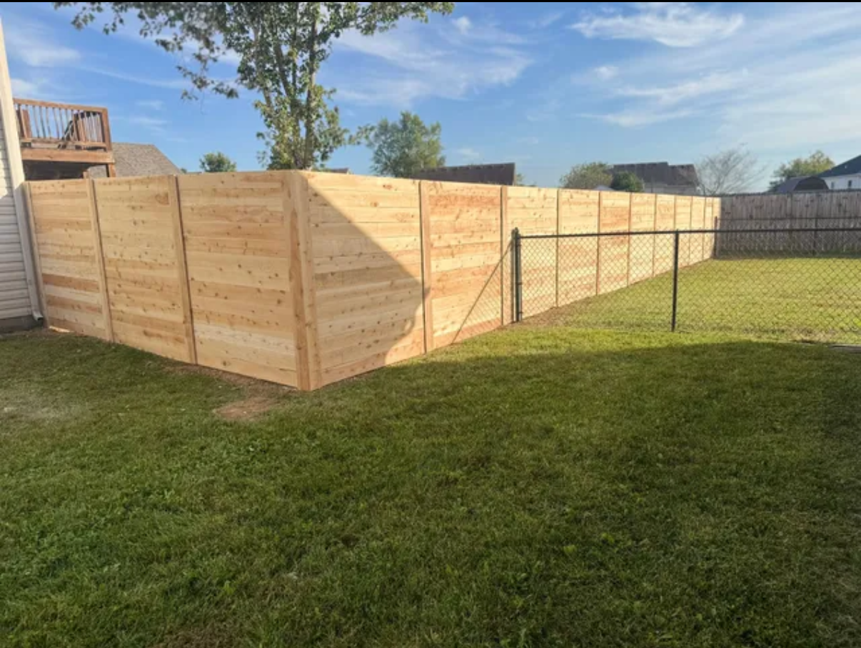 A newly constructed wooden privacy fence stands in a grassy backyard next to an existing chain-link fence.