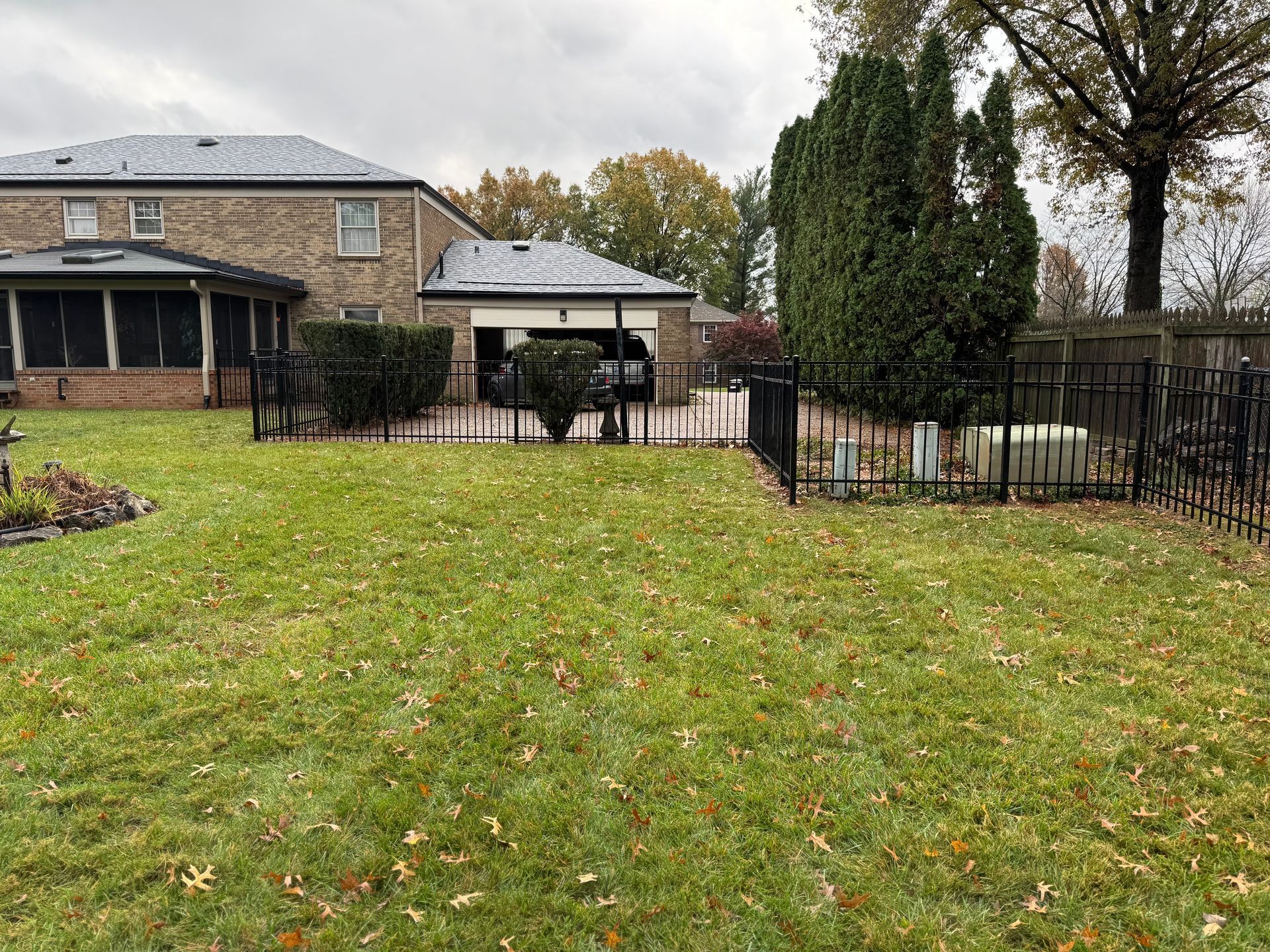 Backyard lawn with black fence, stone house, patio, and trees under cloudy sky