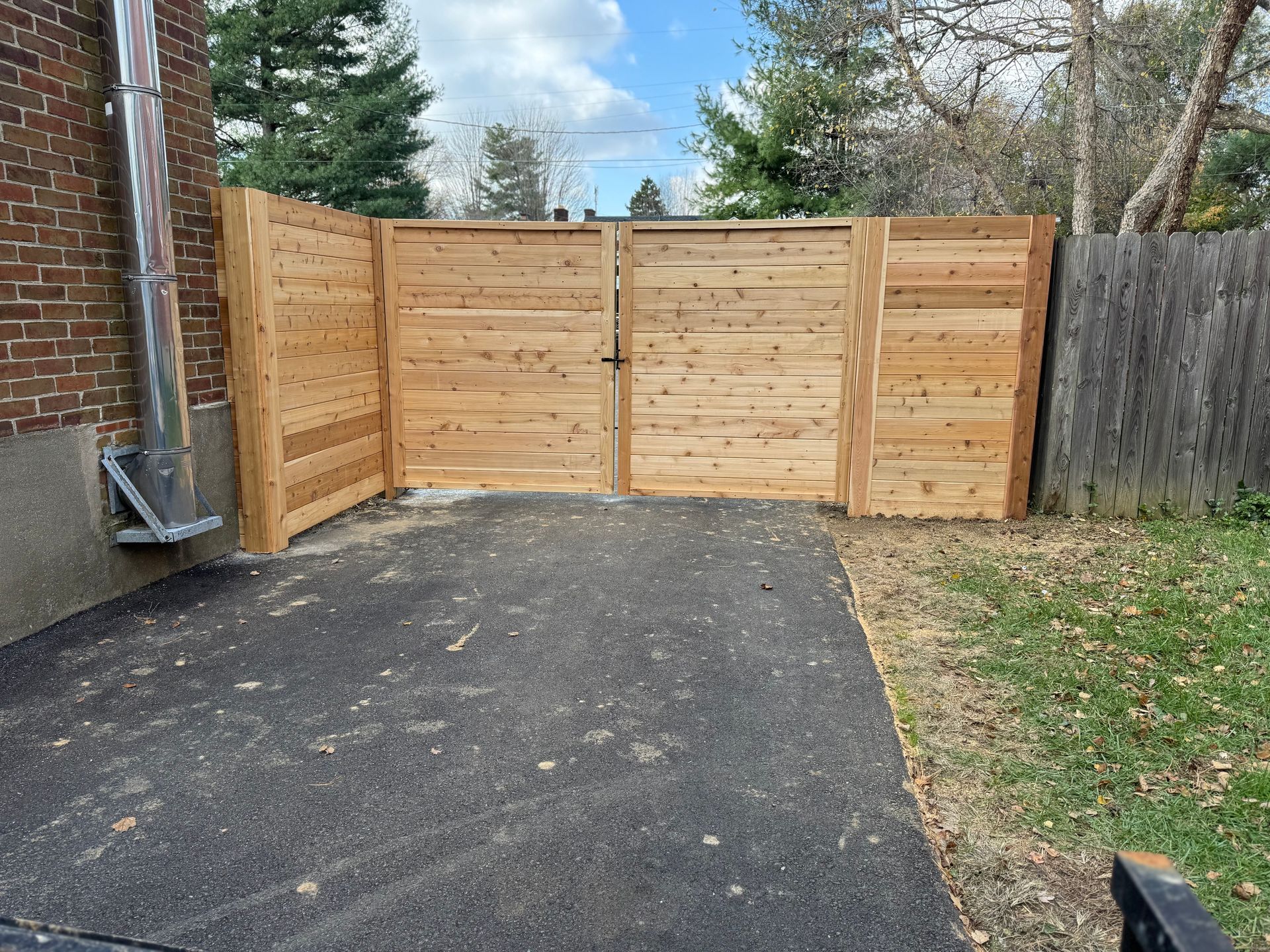 Wooden driveway gate in a fenced yard beside a brick house, with asphalt pavement and grass around it