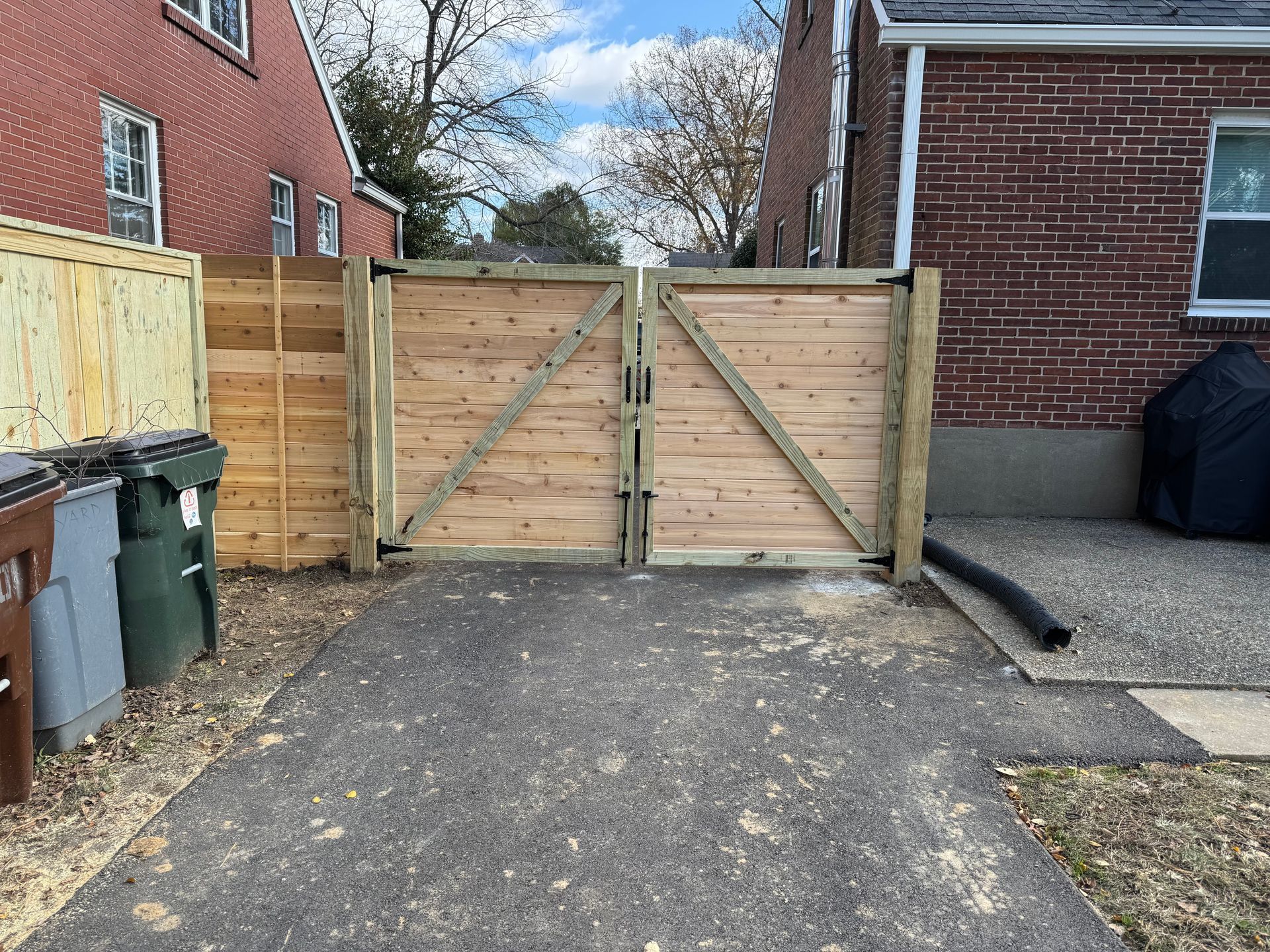 Wooden driveway gate between brick buildings beside paved parking area with trash bins