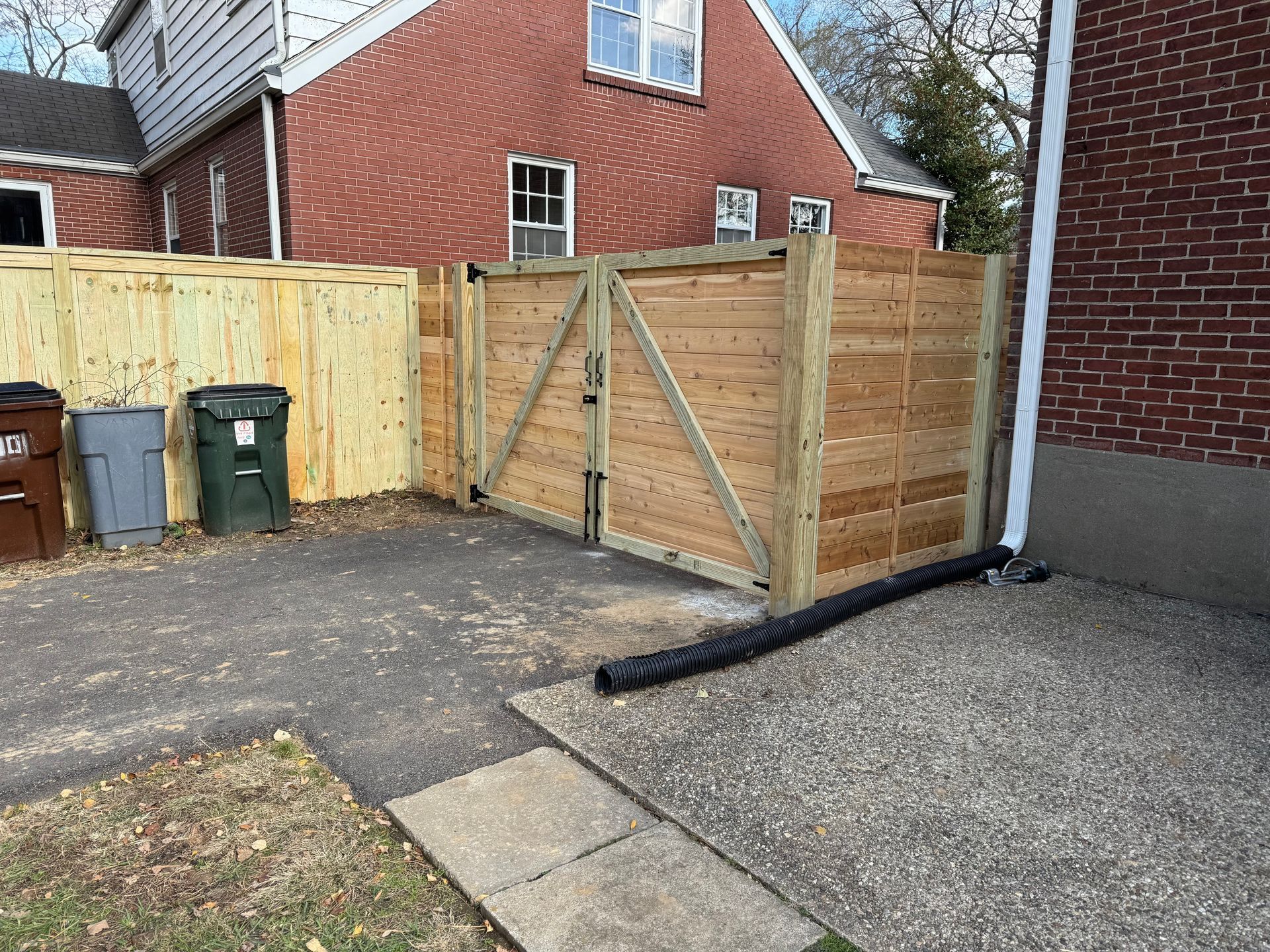 Wooden driveway gate beside a red brick house, with trash bins and a gravel path.