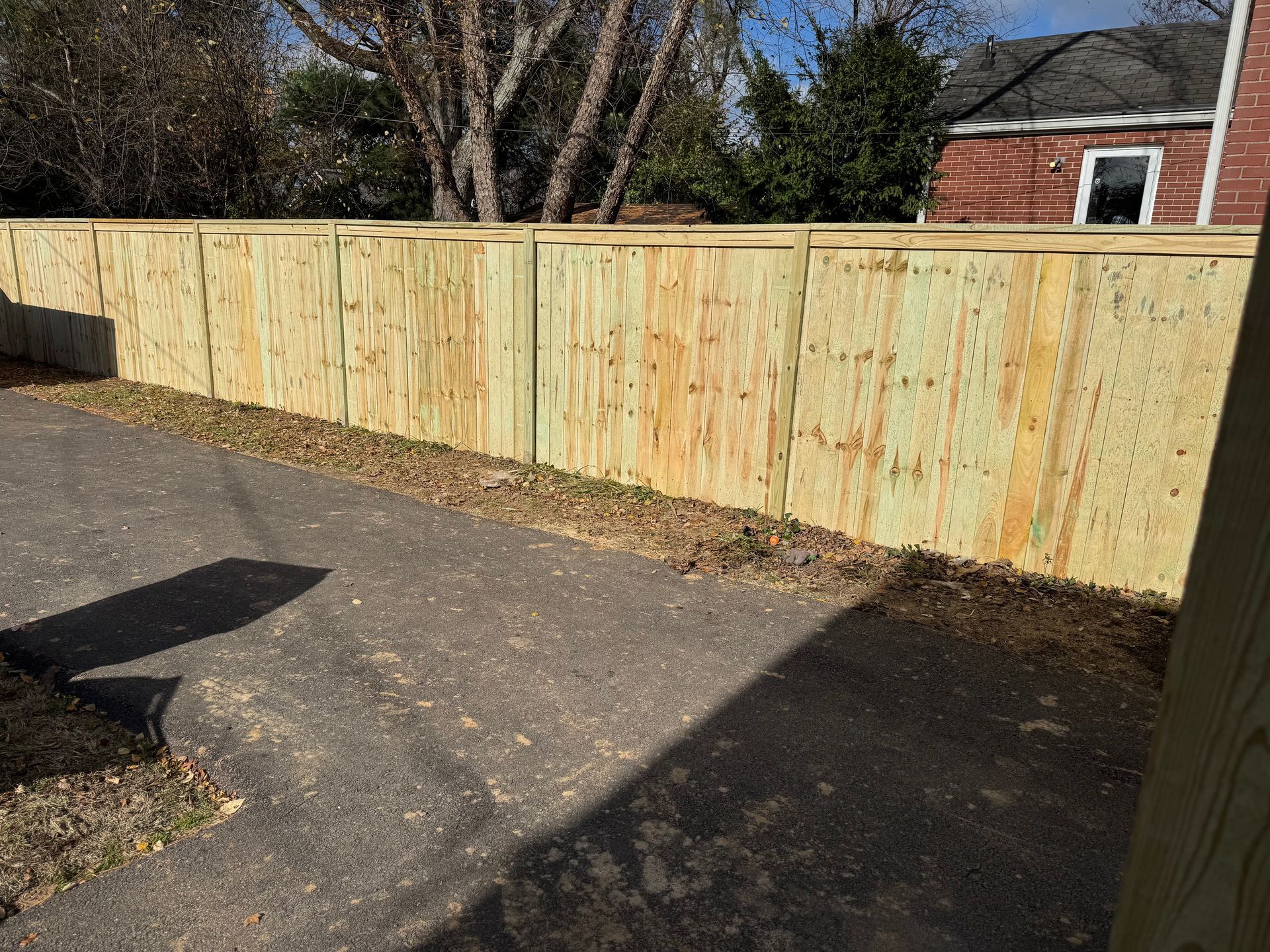 New wooden fence along a paved driveway beside a brick house and trees