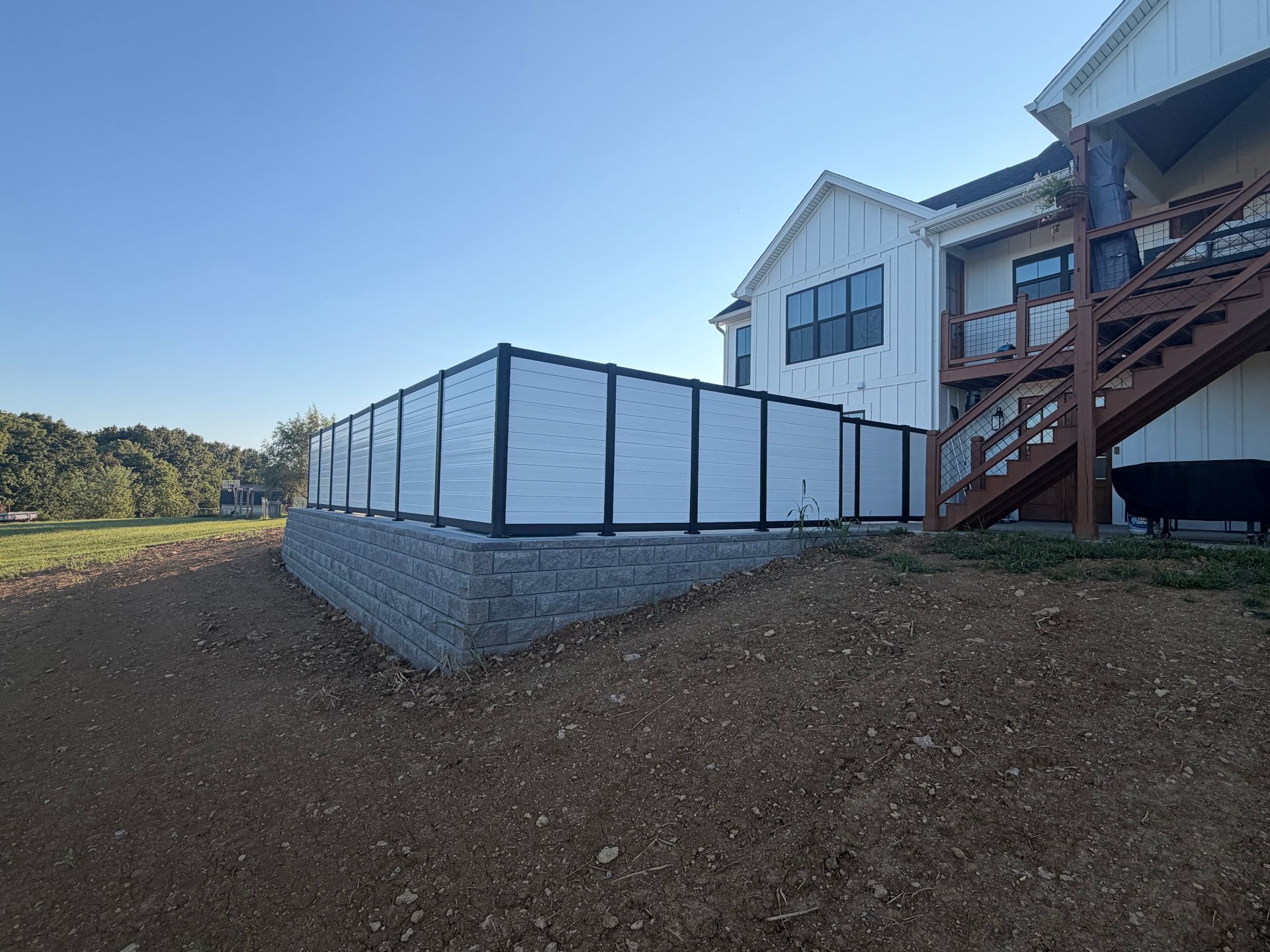 White house with raised deck and staircase beside a fenced retaining wall on a grassy hillside.