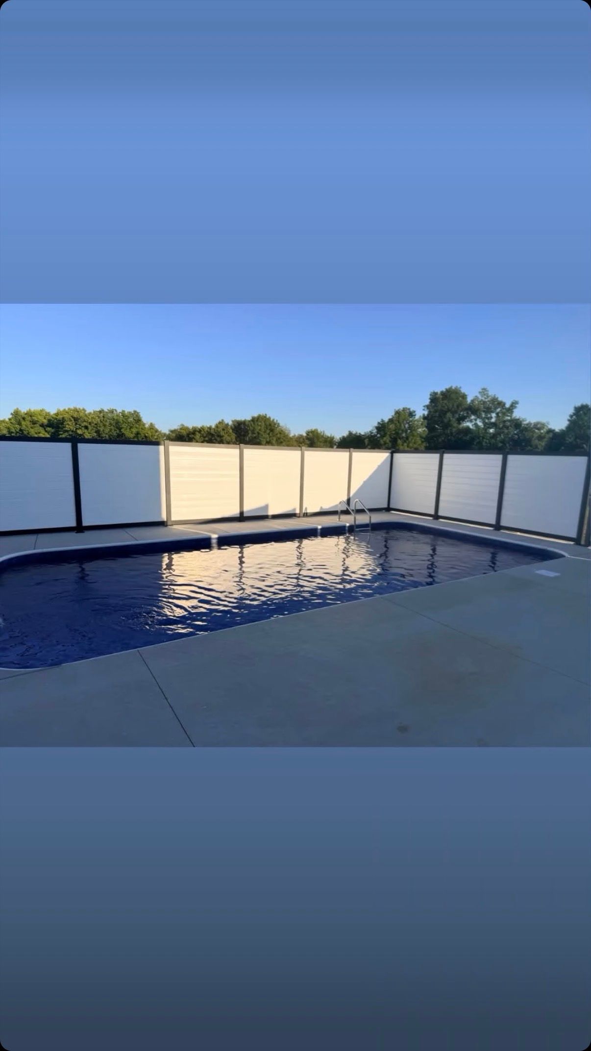 Blue swimming pool at sunset, with white privacy fence and trees in the background.