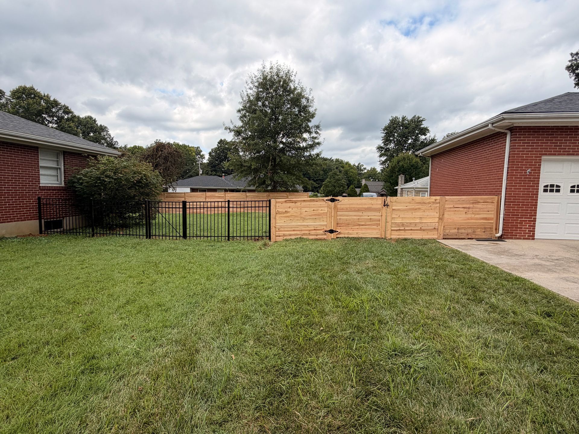 Suburban front yard with a grassy lawn, wooden fence, and brick houses under a cloudy sky