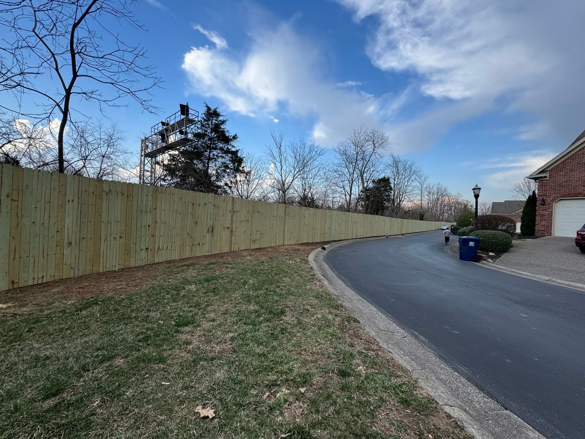 Curved residential driveway beside a wooden fence, with a brick house and blue recycling bins.