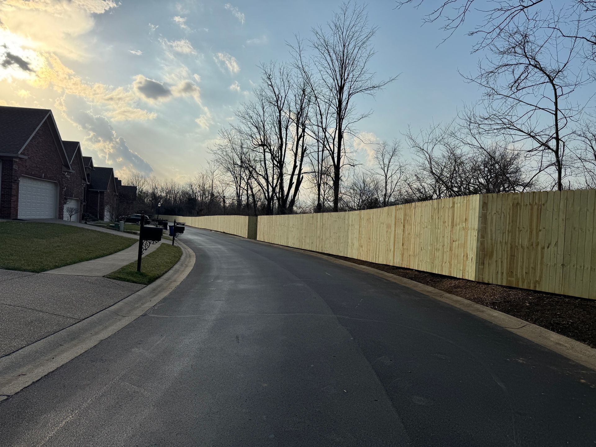 Curving residential street at dusk with houses on the left, tall fence on the right, and bare trees ahead