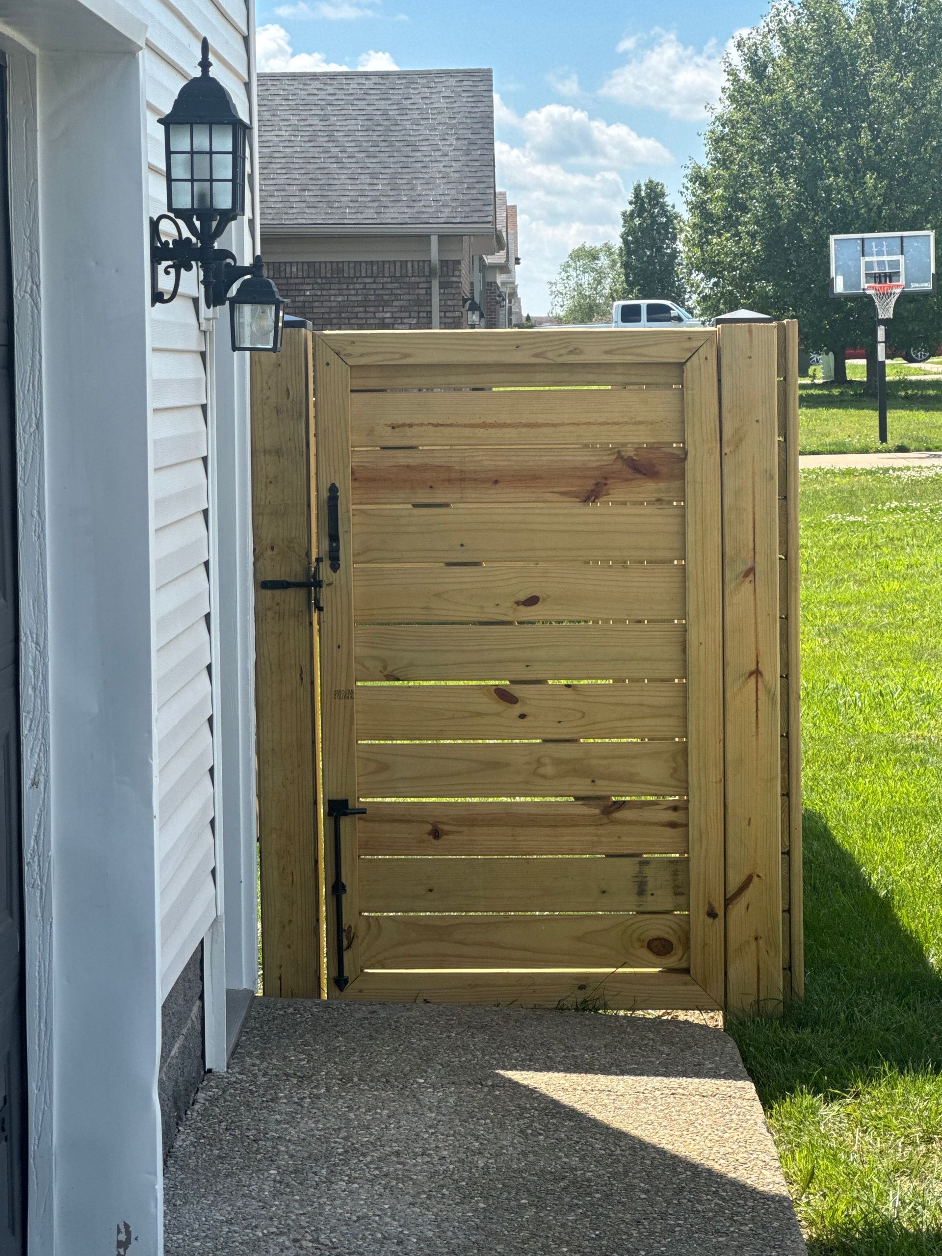 A light-colored wooden gate stands between the corner of a white house and a grassy yard with a basketball hoop.