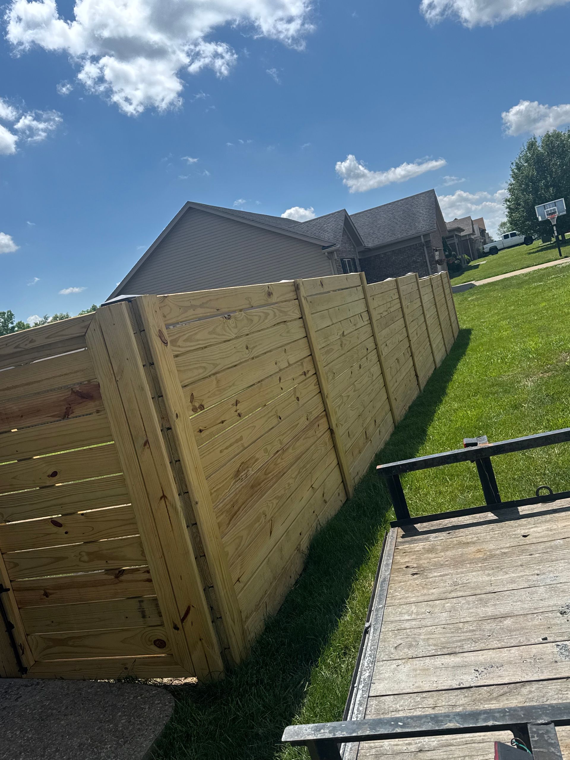Wooden privacy fence beside a grassy yard under a bright blue, cloudy sky.