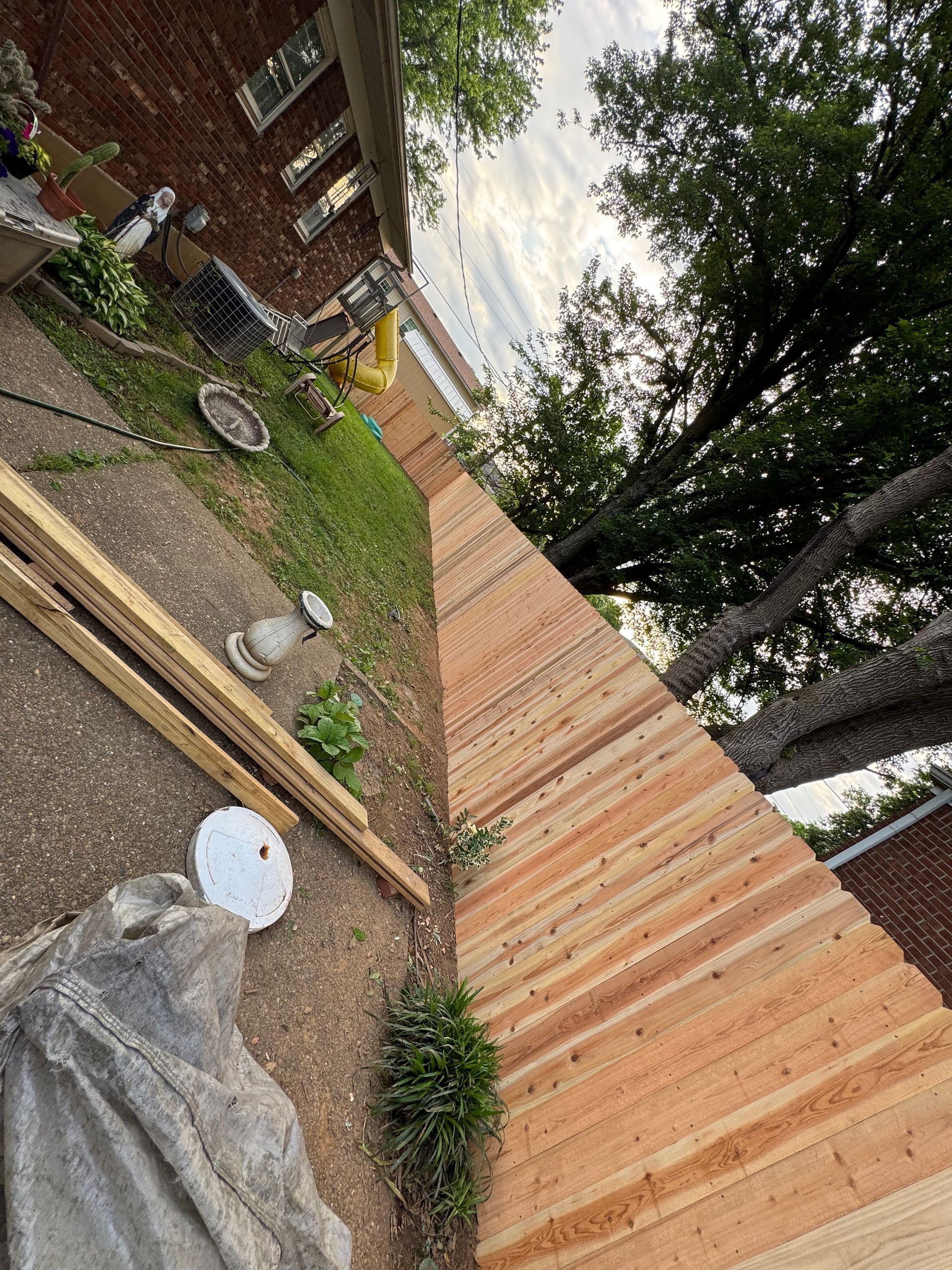 A newly installed wooden fence runs along the side of a brick house, next to a lawn with a bird bath and scattered lumber.