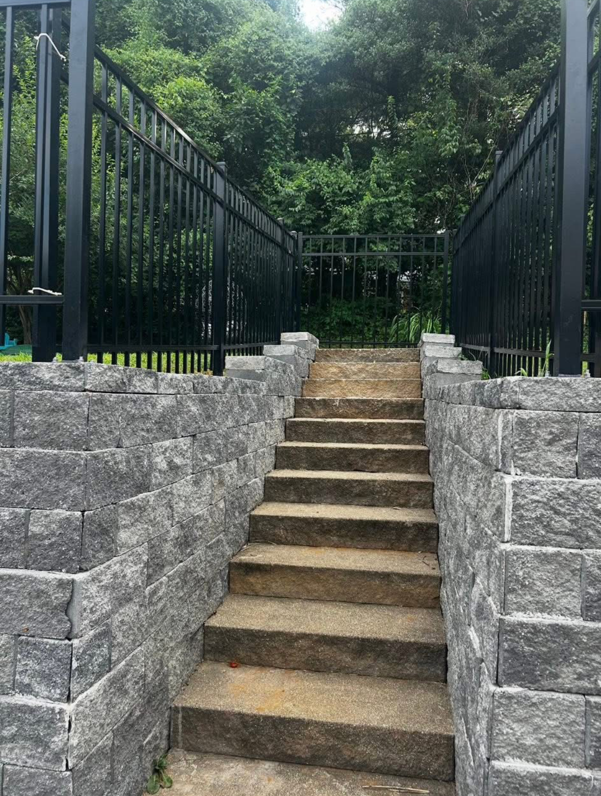 Stone stairs between gray retaining walls, leading up to a black metal fence and green trees above.