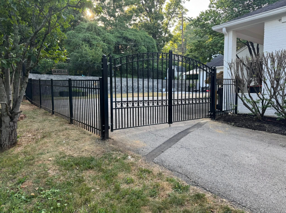 Black metal gate and fence beside a gravel driveway, with trees and a white house in the background