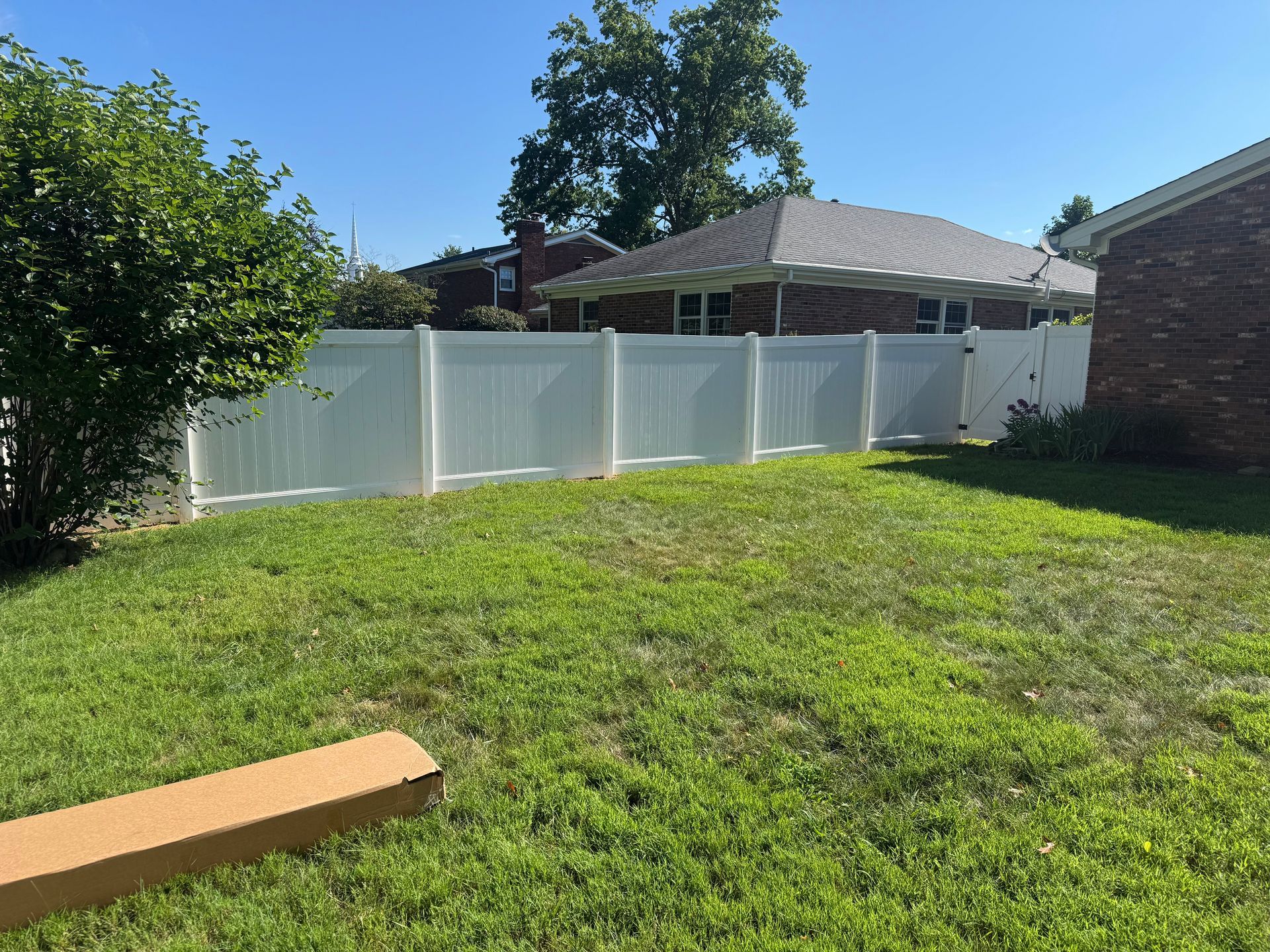 Backyard with white vinyl fence, green grass, and a cardboard box. Buildings and trees in the background.