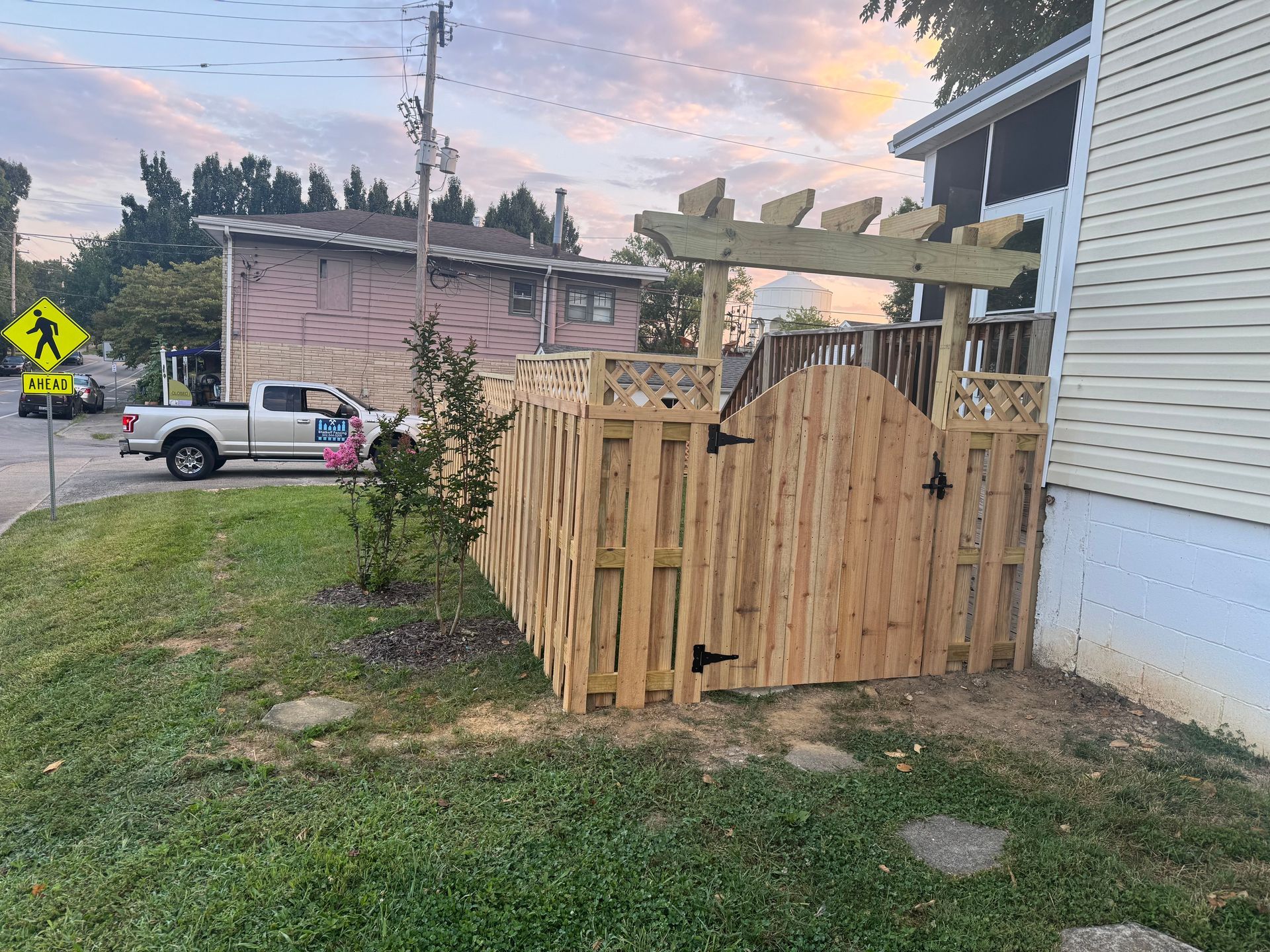 Wooden backyard fence and gate beside a white house, with a pickup truck and street in the background at dusk