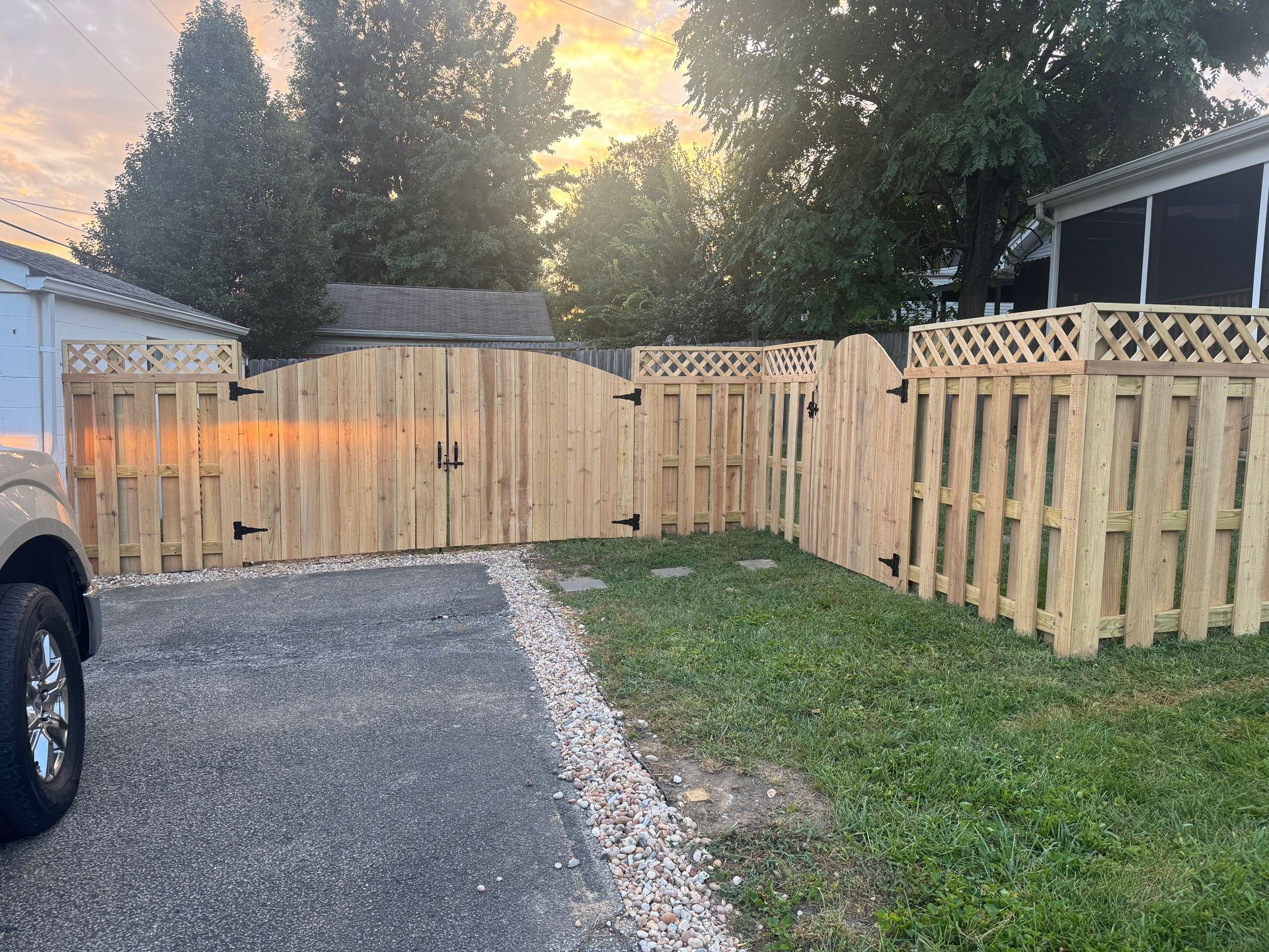 Gravel driveway beside a wooden fence and gate, with a parked truck and trees at sunset.