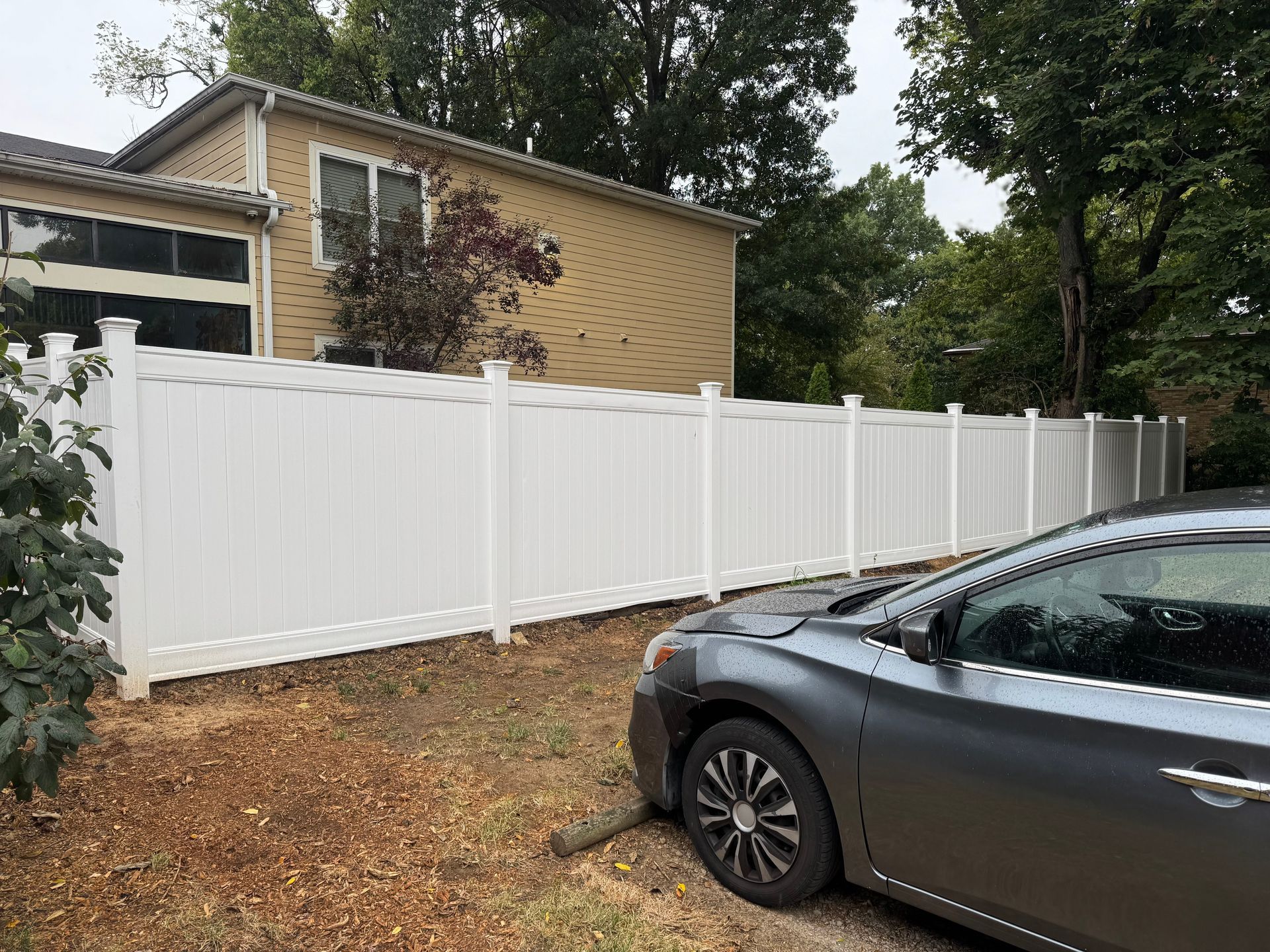 White vinyl fence in a yard with a gray car parked near it. Beige house in the background.