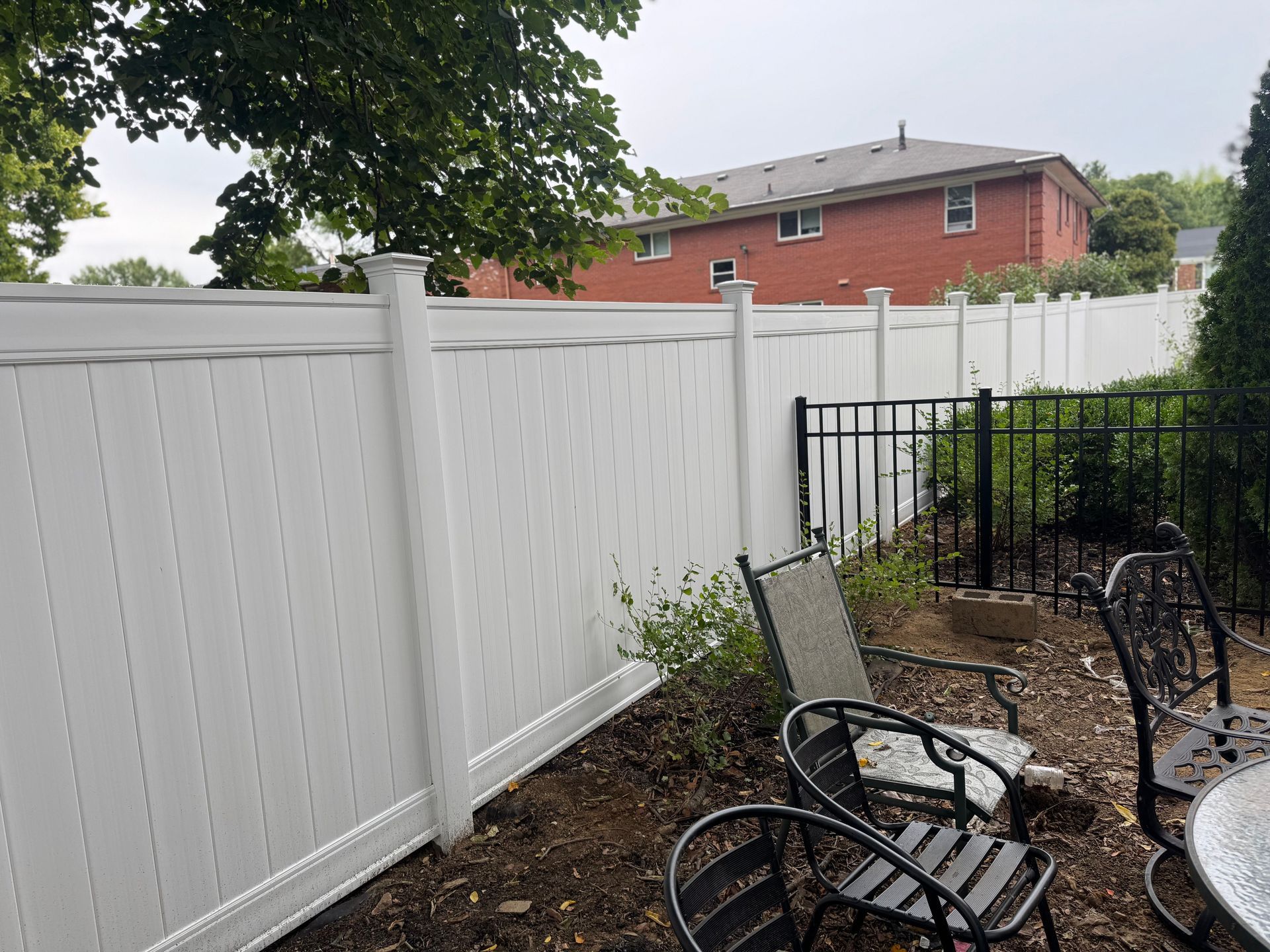 White vinyl fence bordering a yard with patio furniture. Red brick house in the background.