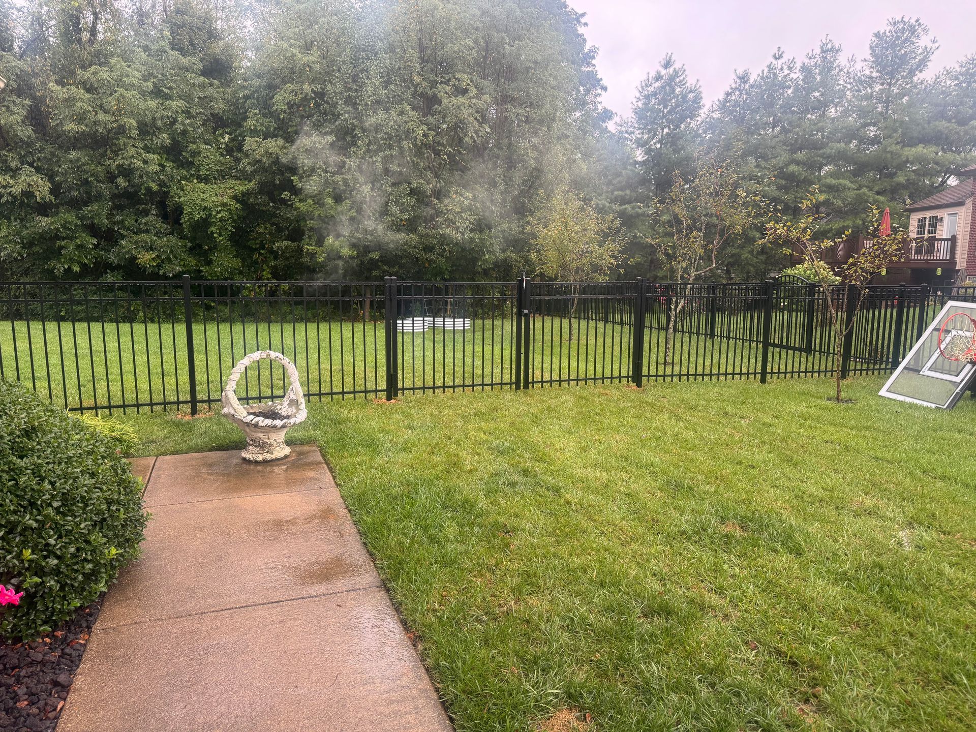 Backyard lawn with stone path, white chair, black fence, and a soccer goal near trees.