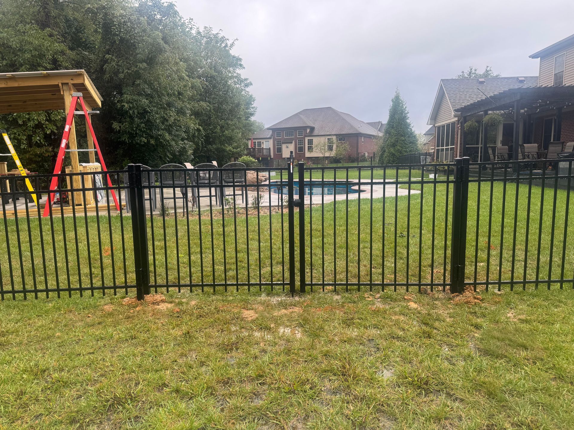 Backyard with black metal fence, playset, grass lawn, and houses under cloudy sky