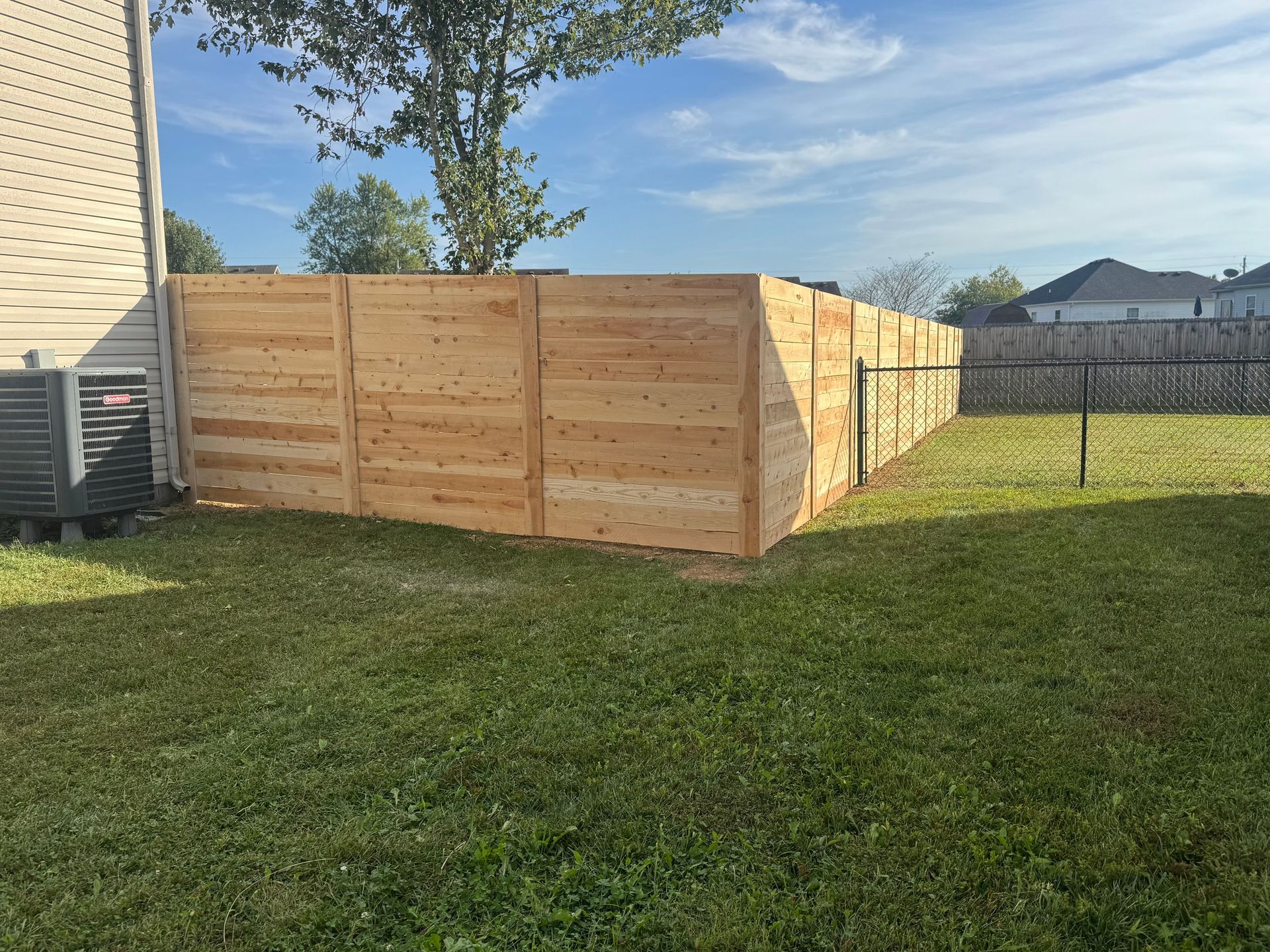 Wooden privacy fence around a grassy backyard beside a house, with a tree and neighboring homes in view
