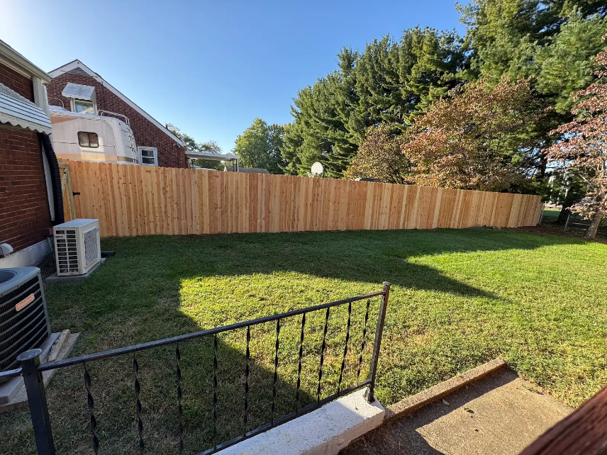 Backyard lawn with wooden fence, brick house, trees, and a small patio railing in the foreground