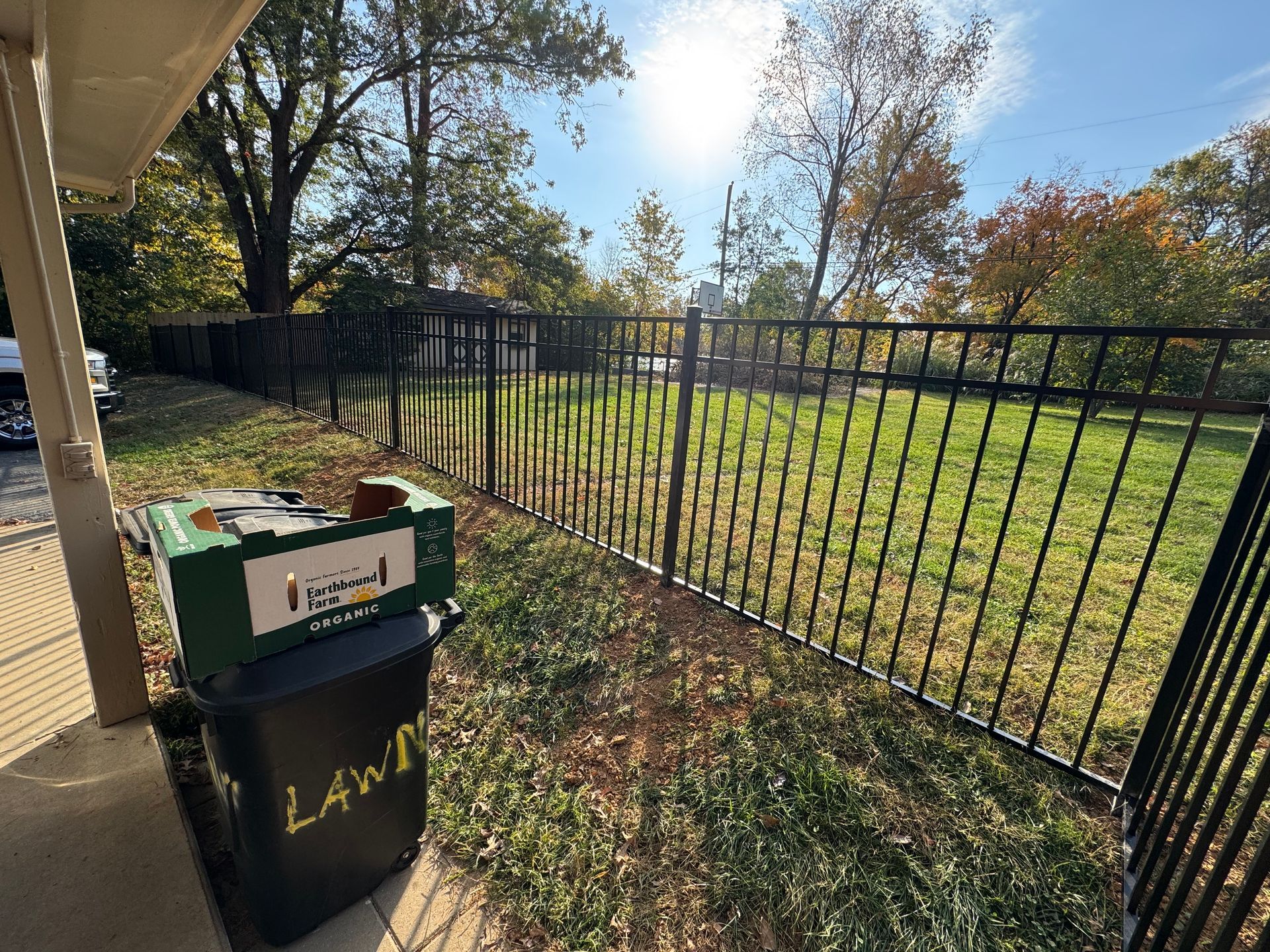 Black trash bin and cardboard box beside a fenced grassy yard on a sunny day
