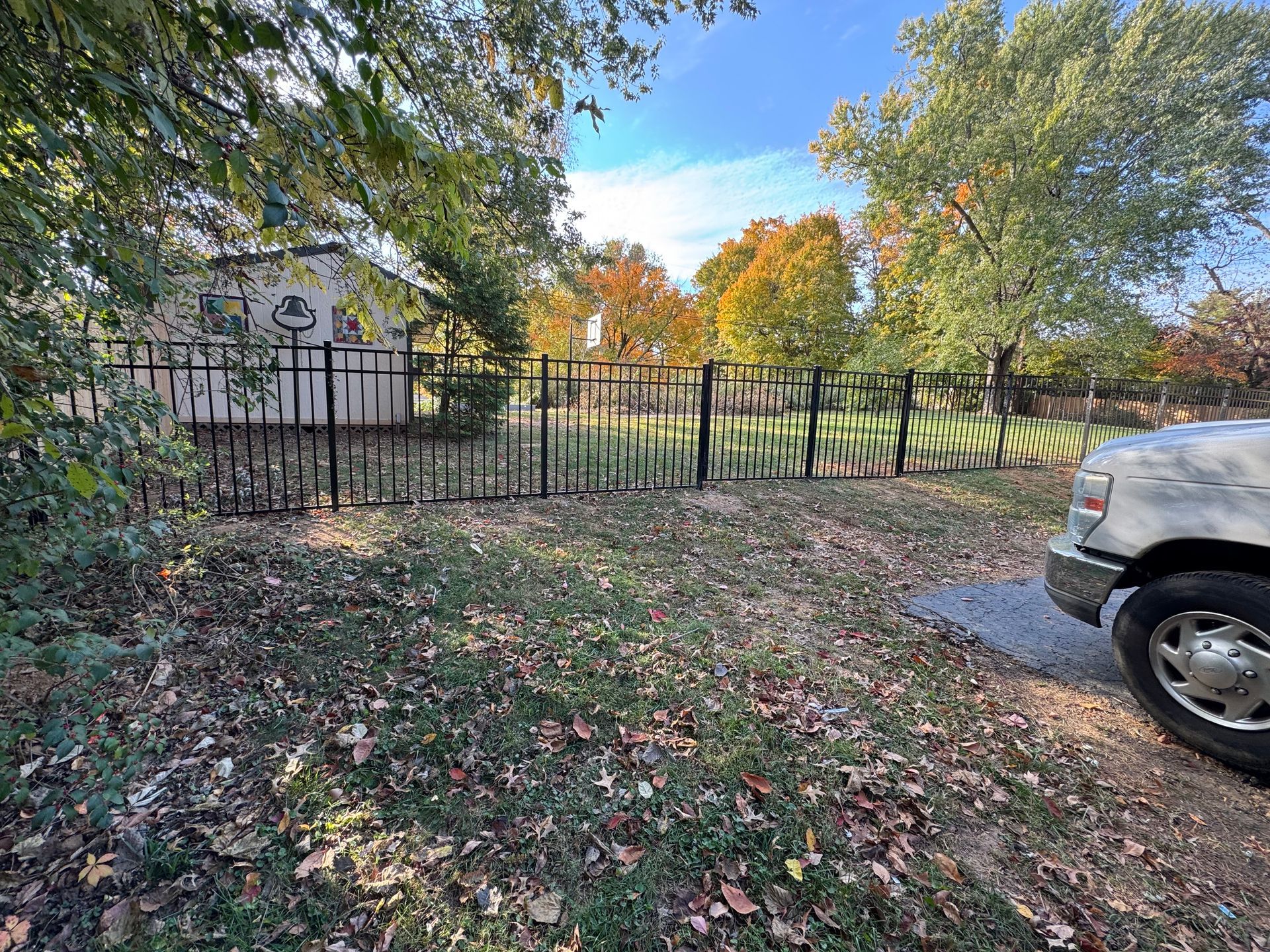Leaf-covered yard with a black metal fence, trees with autumn foliage, and part of a white truck at right.