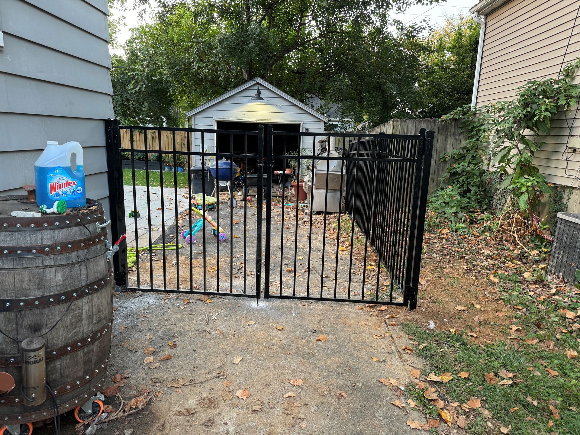 Backyard gate opening to a cluttered driveway and small shed, with a barrel and leaves along the fence