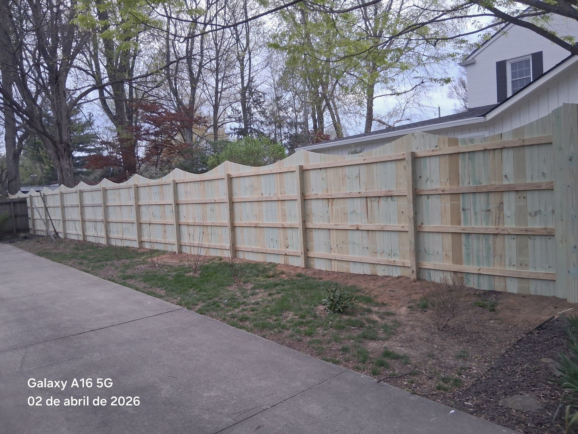 A new, light-colored wooden scalloped-top fence runs along a concrete driveway next to a house.