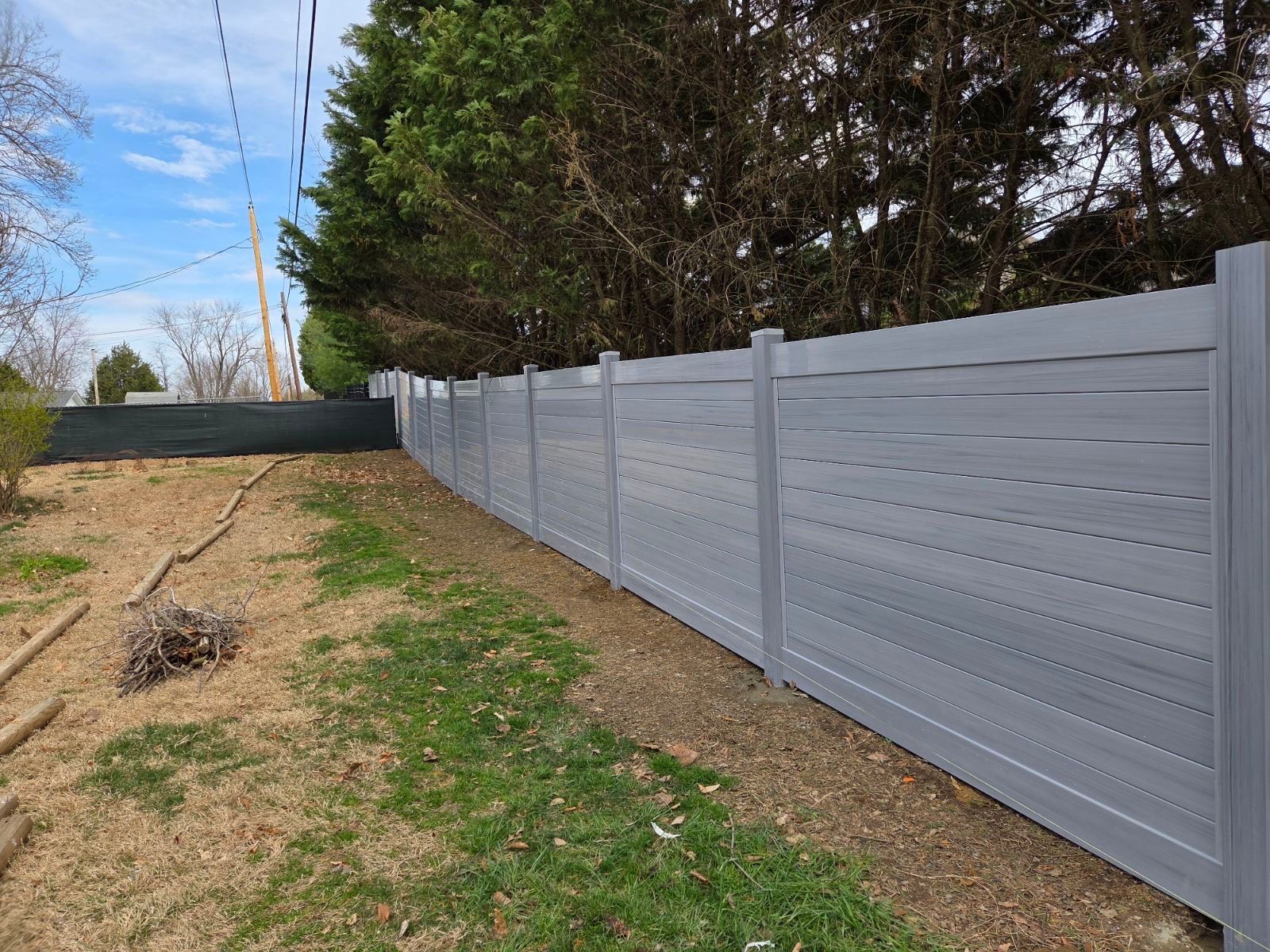 Gray horizontal slat fence in a yard with sparse grass and trees in the background.