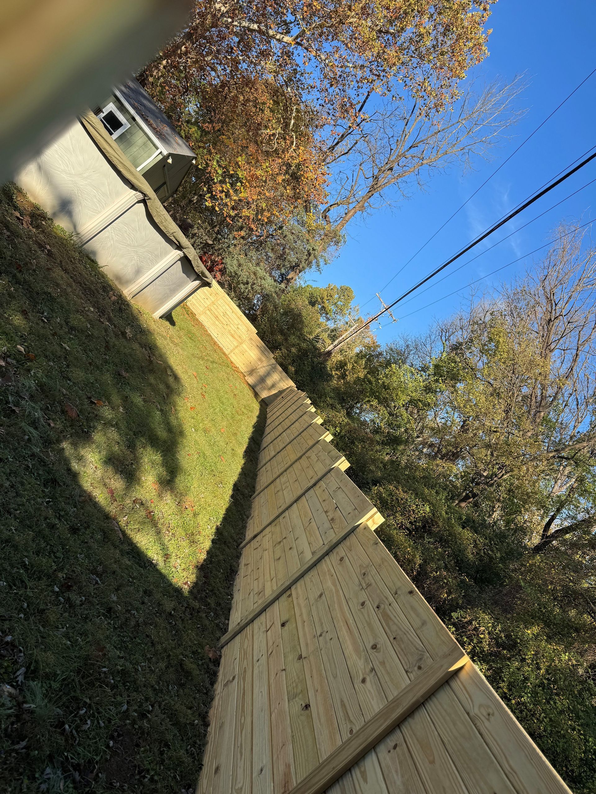 Tilted view of a wooden fence beside a grassy yard, with trees and a blue sky behind it.