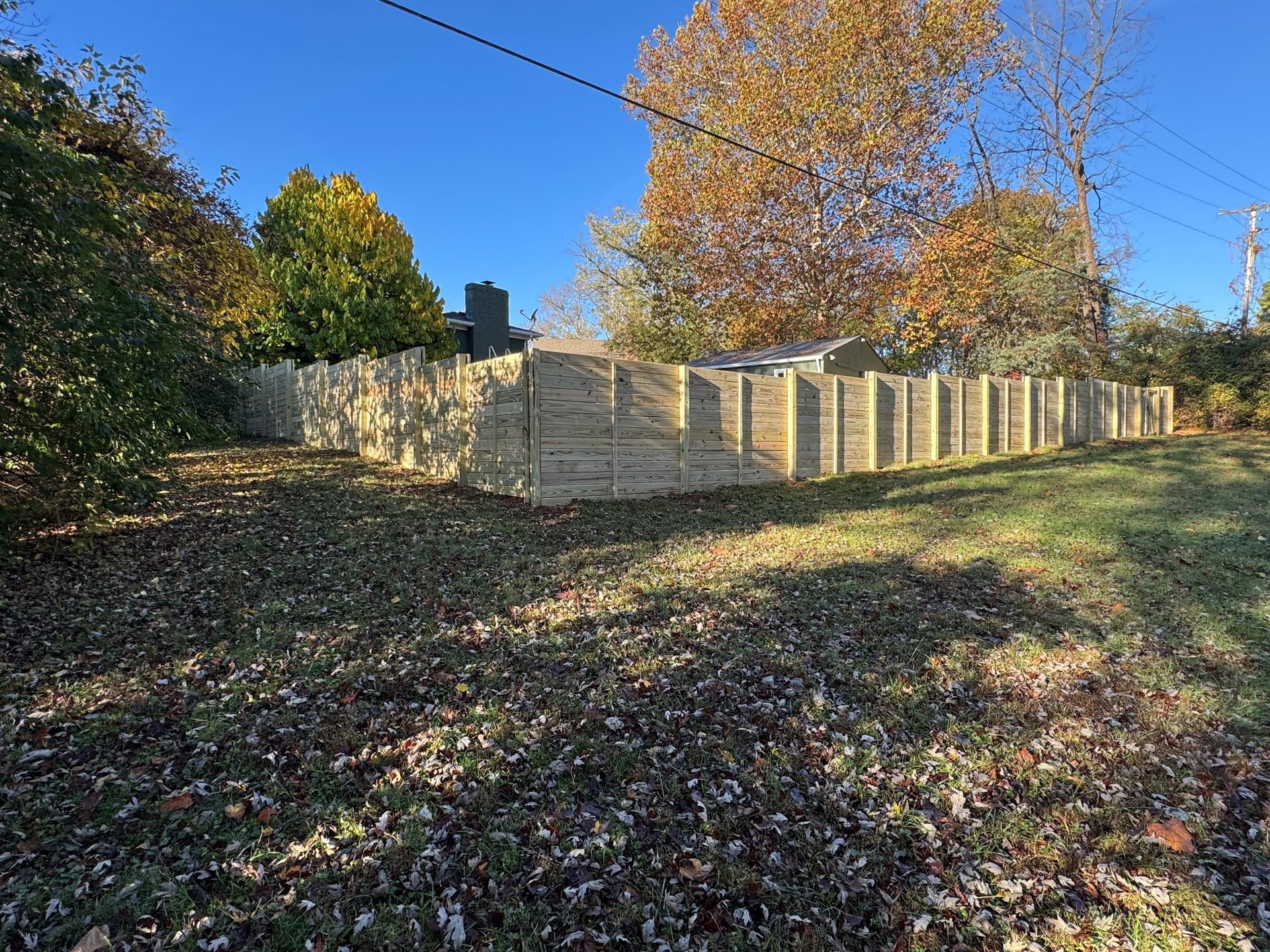 Wooden fence along a leaf-covered grassy slope under blue sky and autumn trees