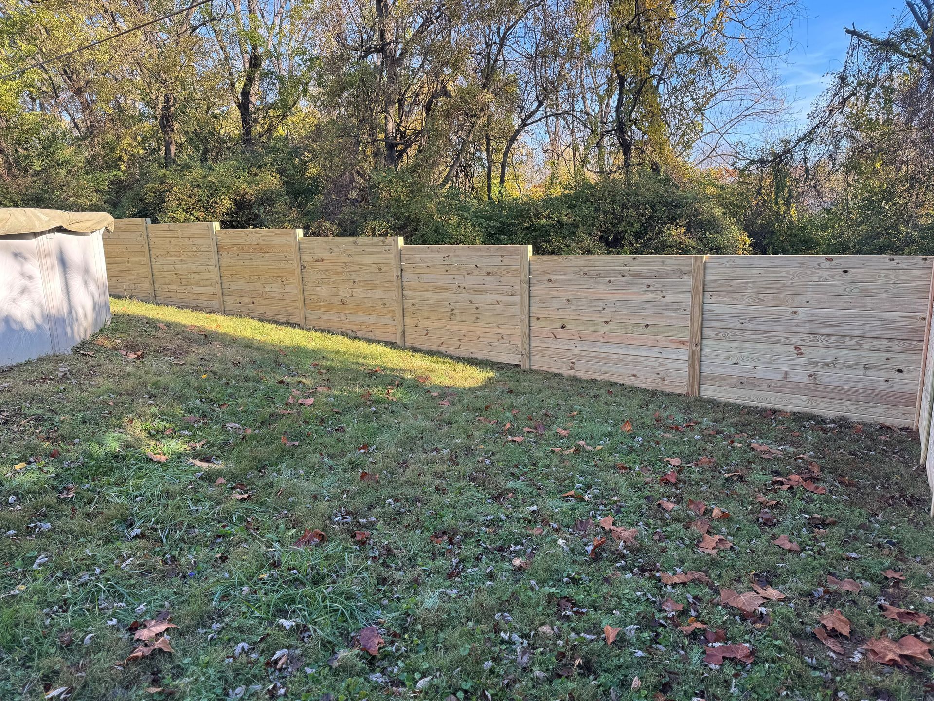 Wooden privacy fence along a grassy backyard with trees and blue sky behind it
