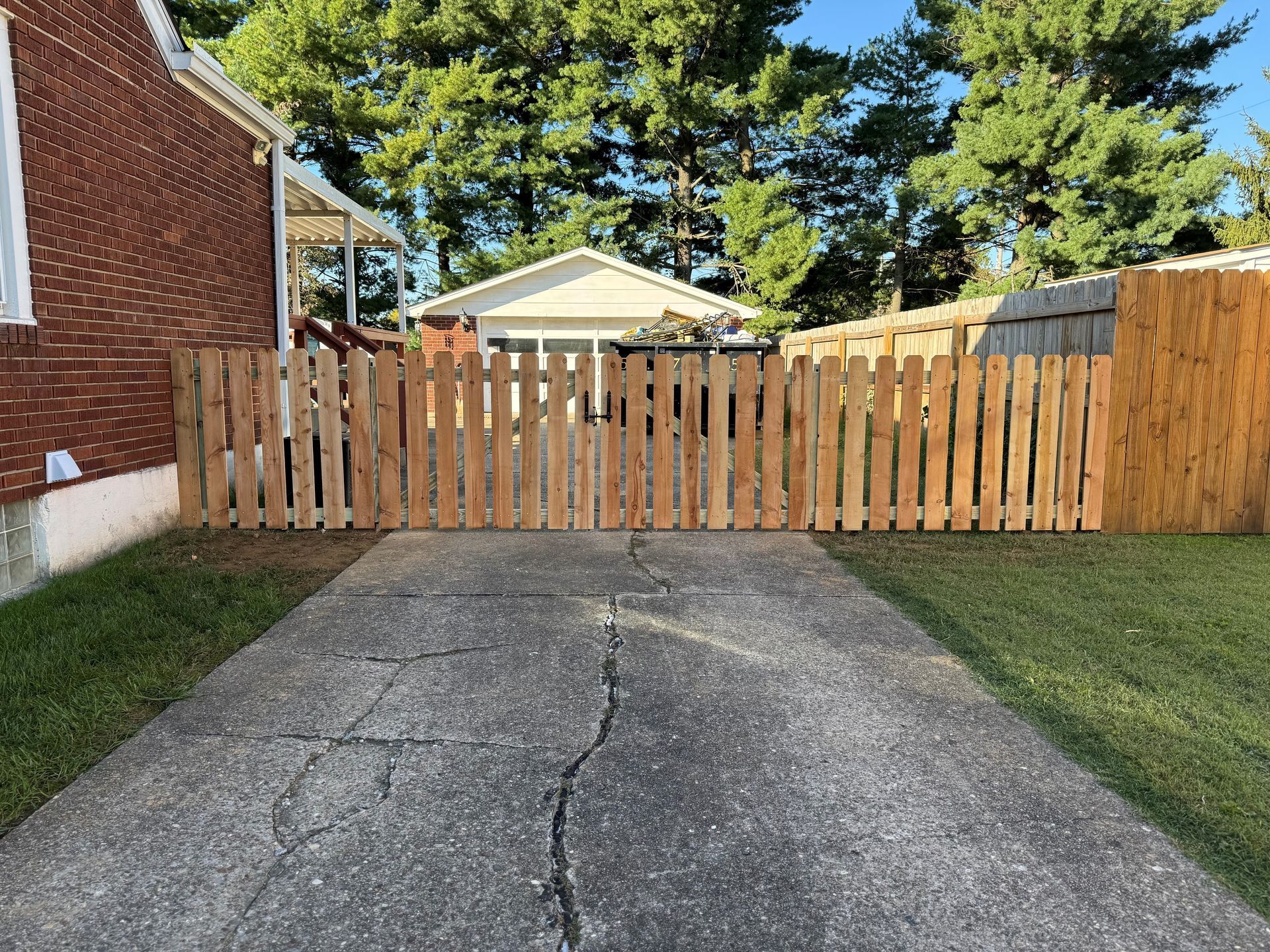 Driveway leading to a wooden fence gate, with a brick house on the left and trees behind.